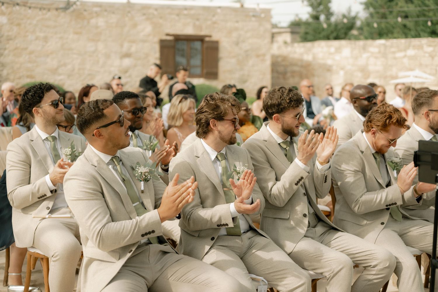 Groomsmen in matching beige suits clapping at an outdoor wedding ceremony.