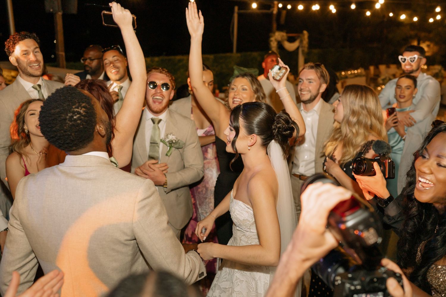 Bride and groom celebrate joyfully with guests dancing at an outdoor wedding reception under string lights.