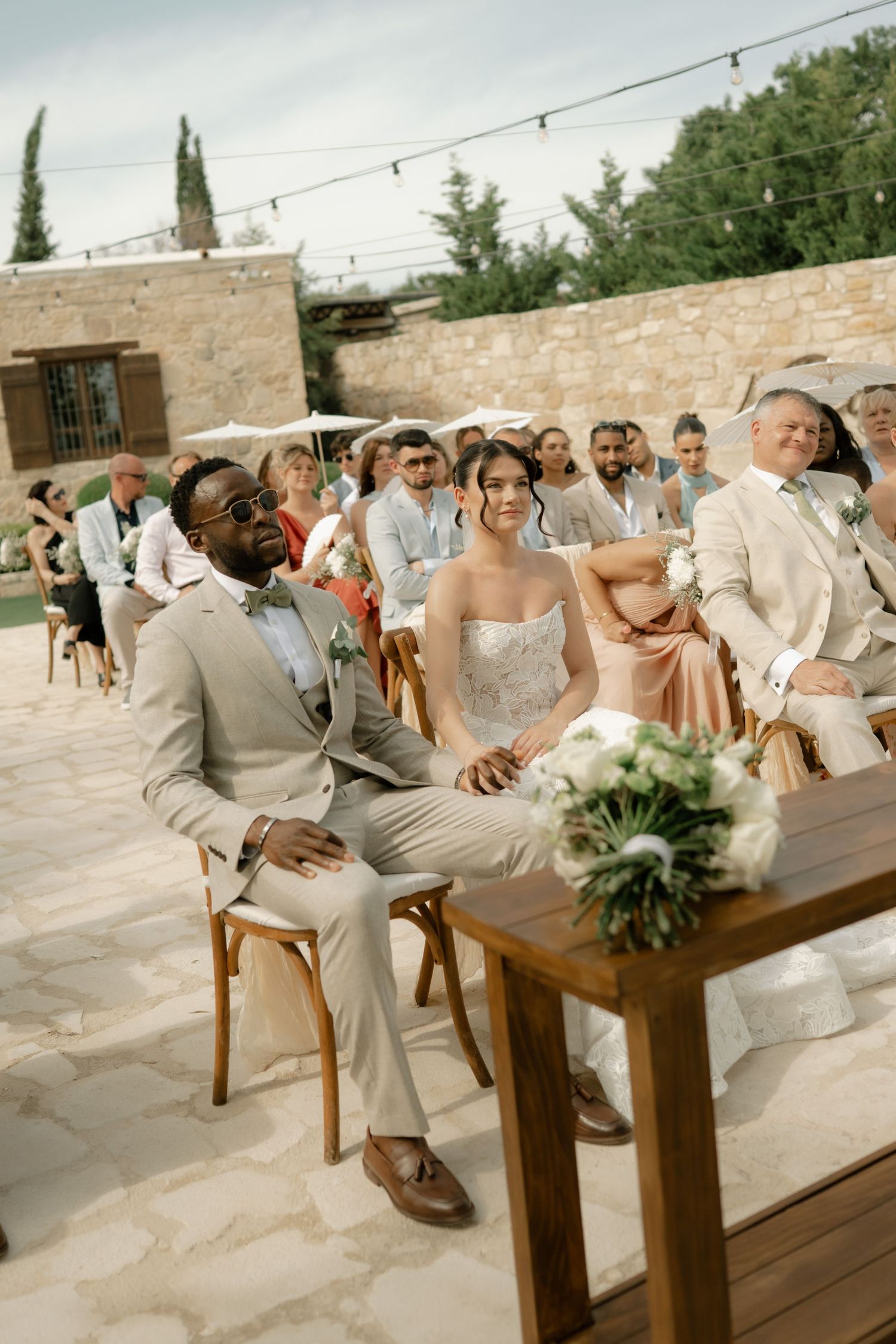 Bride and groom seated at outdoor wedding ceremony with guests in background at a rustic stone venue.
