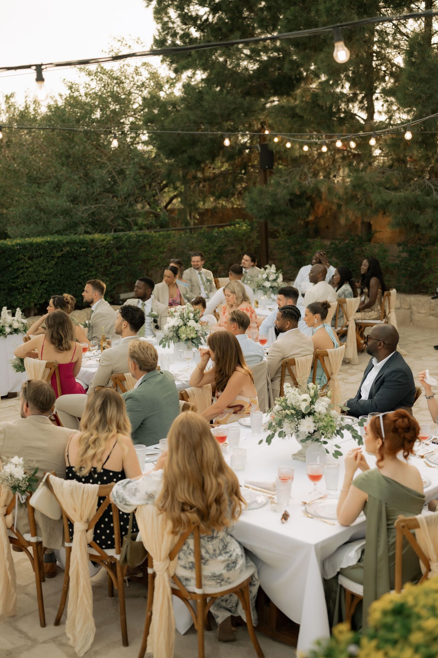 Guests seated at elegant outdoor wedding reception tables with white florals and string lights at dusk.