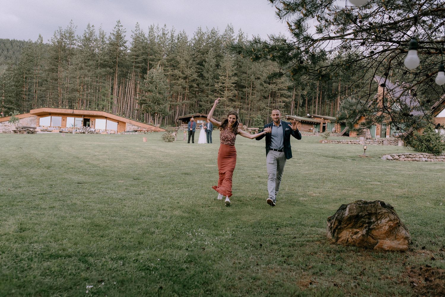 People mingling on a spacious lawn with pine forest and cabins in the background.