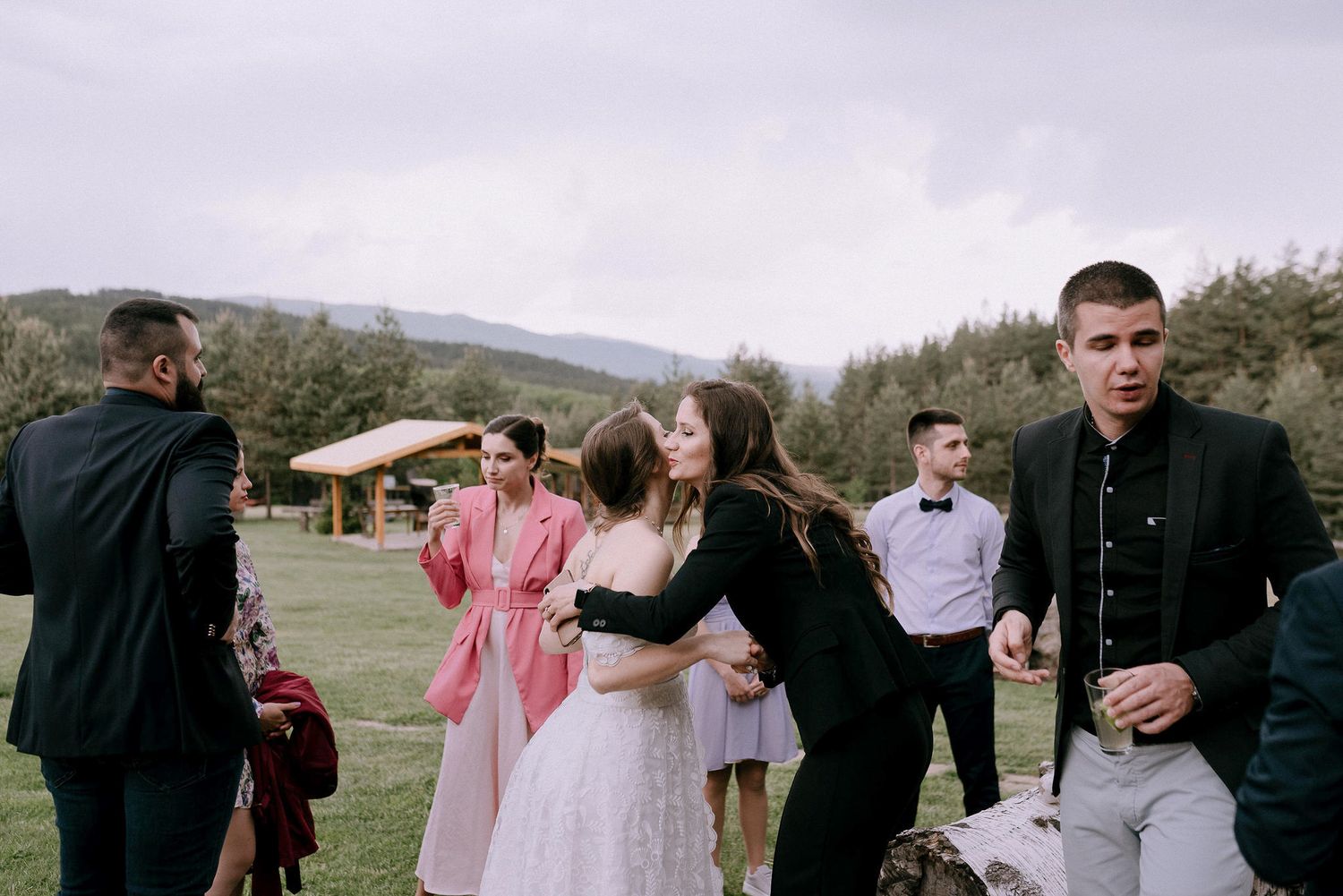 Wedding celebration with guests mingling on a grassy hillside near pine trees.