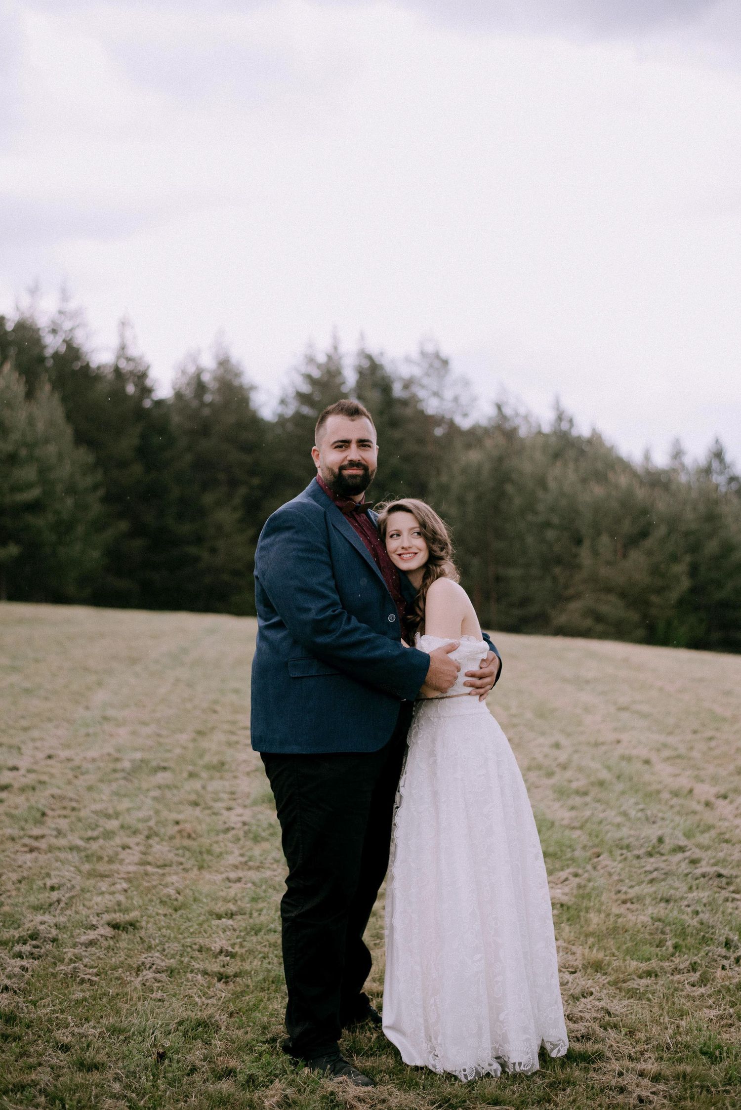 A couple embraces on a scenic hillside with evergreen trees in the background.