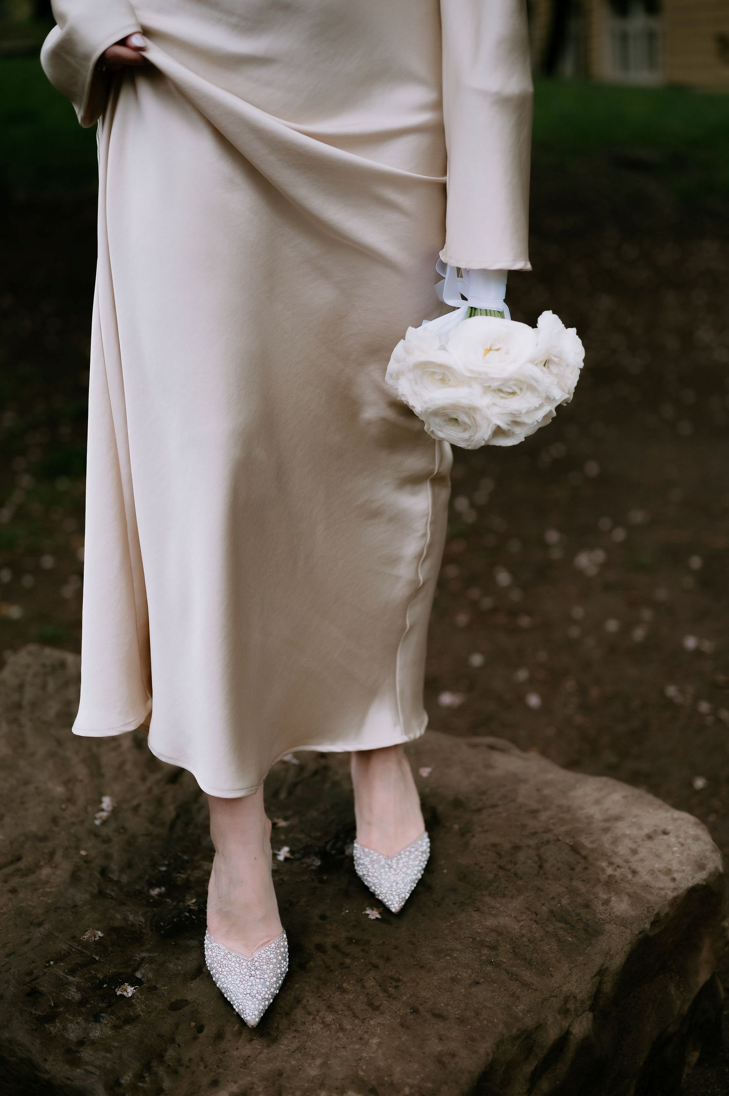 Close-up of sparkly white bridal shoes and dress hem on a natural stone surface.