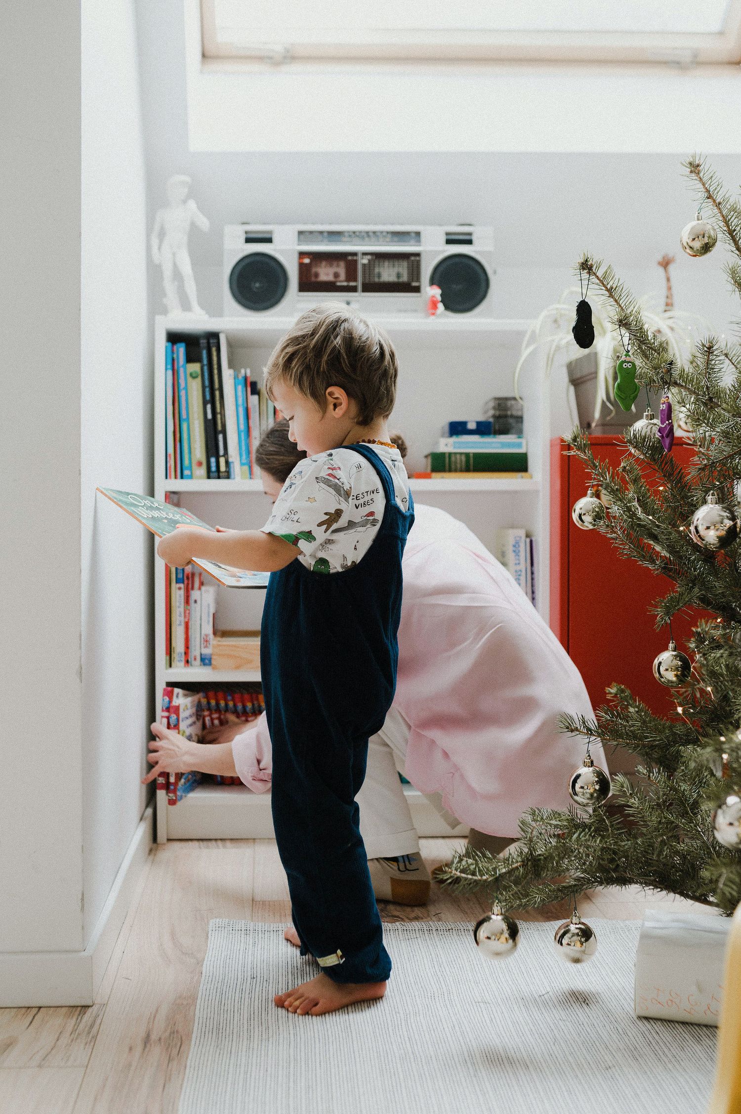Small child in overalls stands reading a book near a decorated Christmas tree and bookshelf with a vintage boombox.