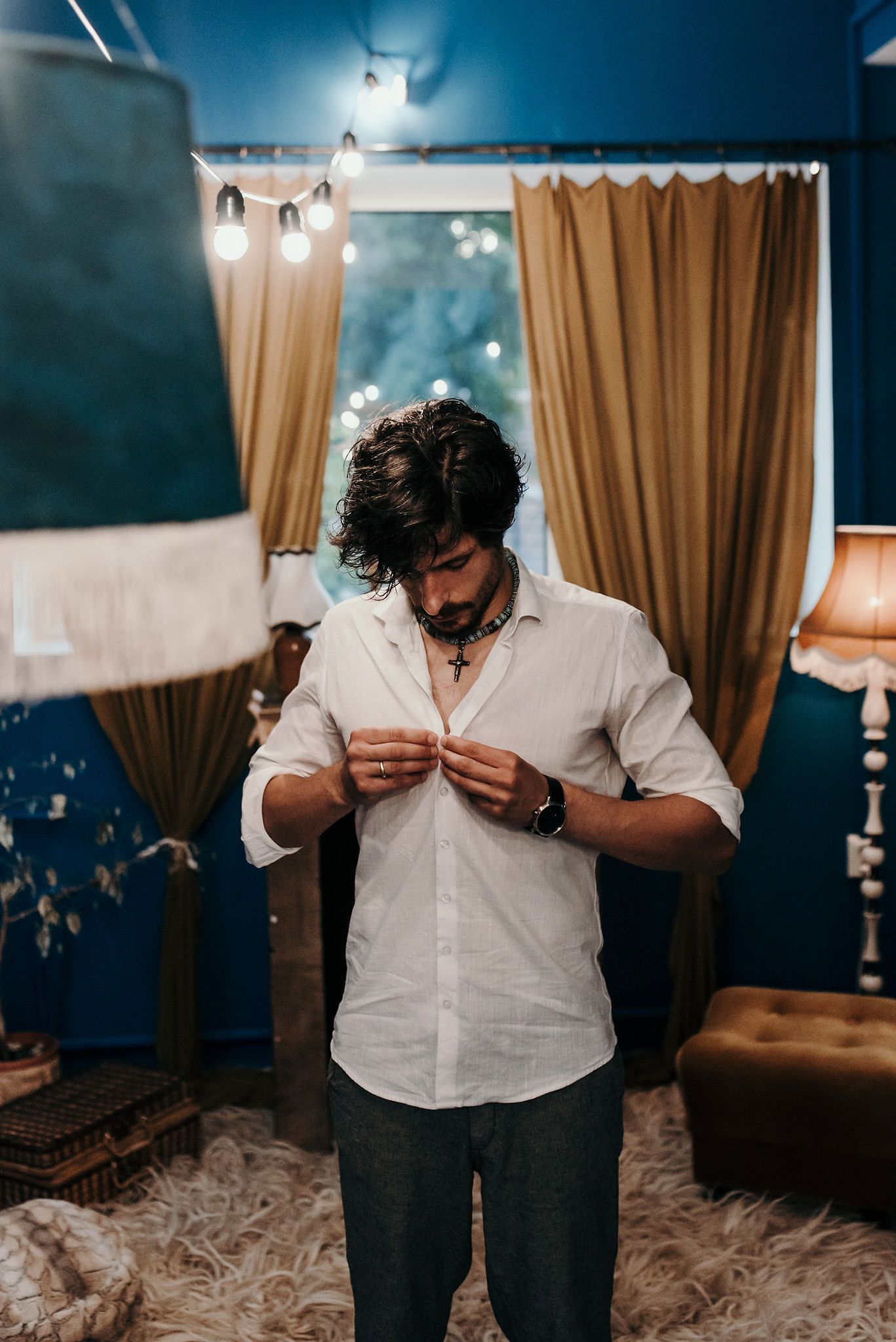 Person buttoning a white dress shirt in a room with blue walls and gold curtains.
