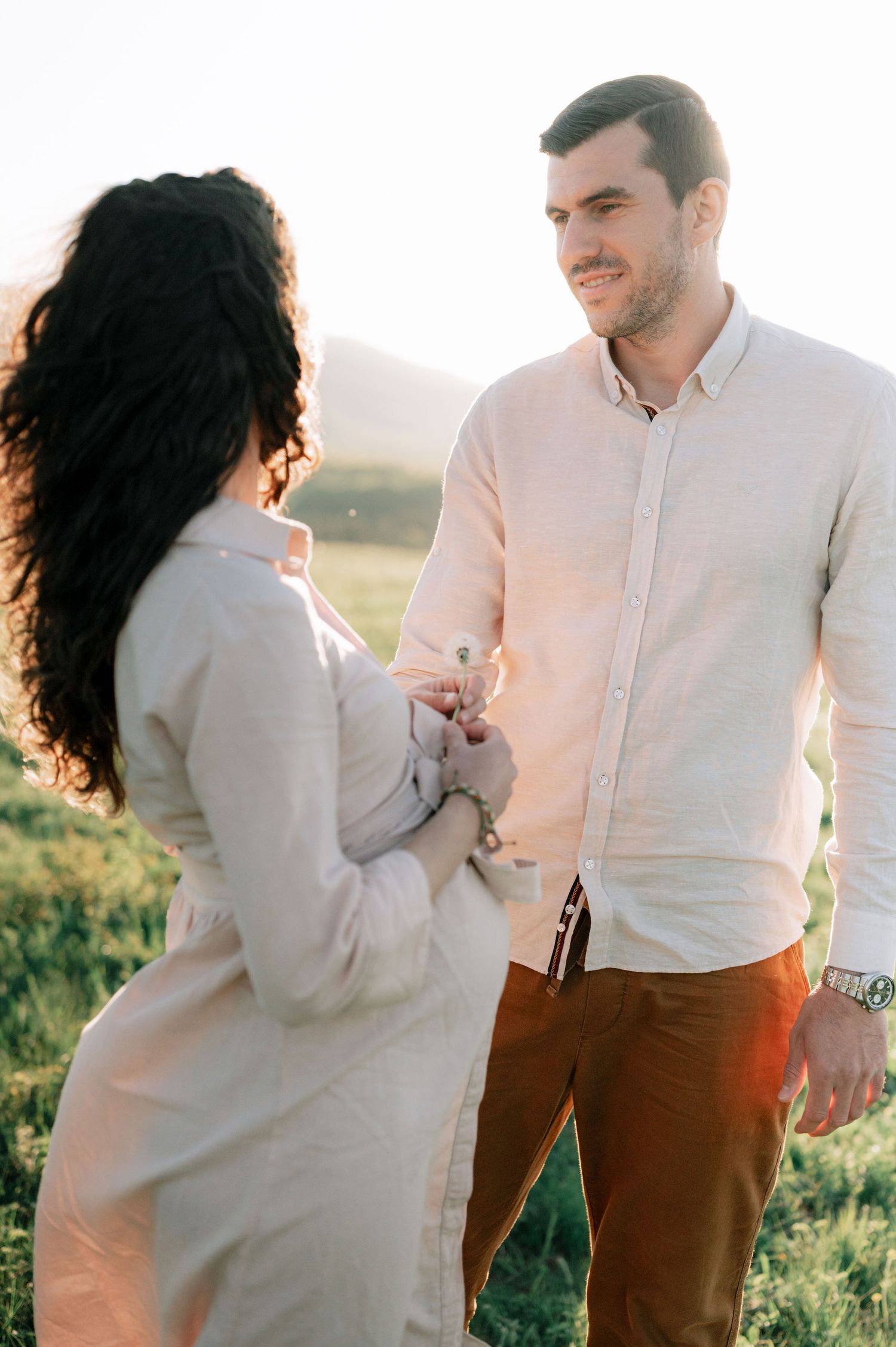 A couple shares intimate moments in a sunlit field while wearing coordinated white and brown outfits.