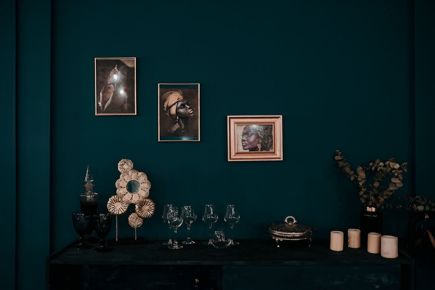 Vintage photographs hang on a dark teal wall above wine glasses and decorative items on a table.