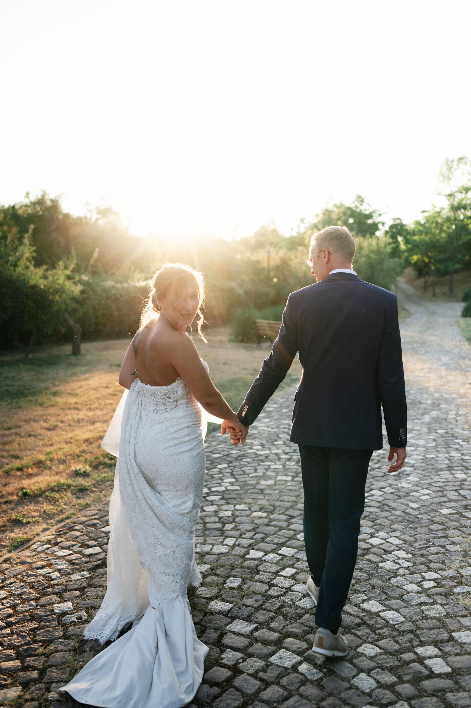 Newlyweds holding hands while walking down a scenic cobblestone path during golden hour.