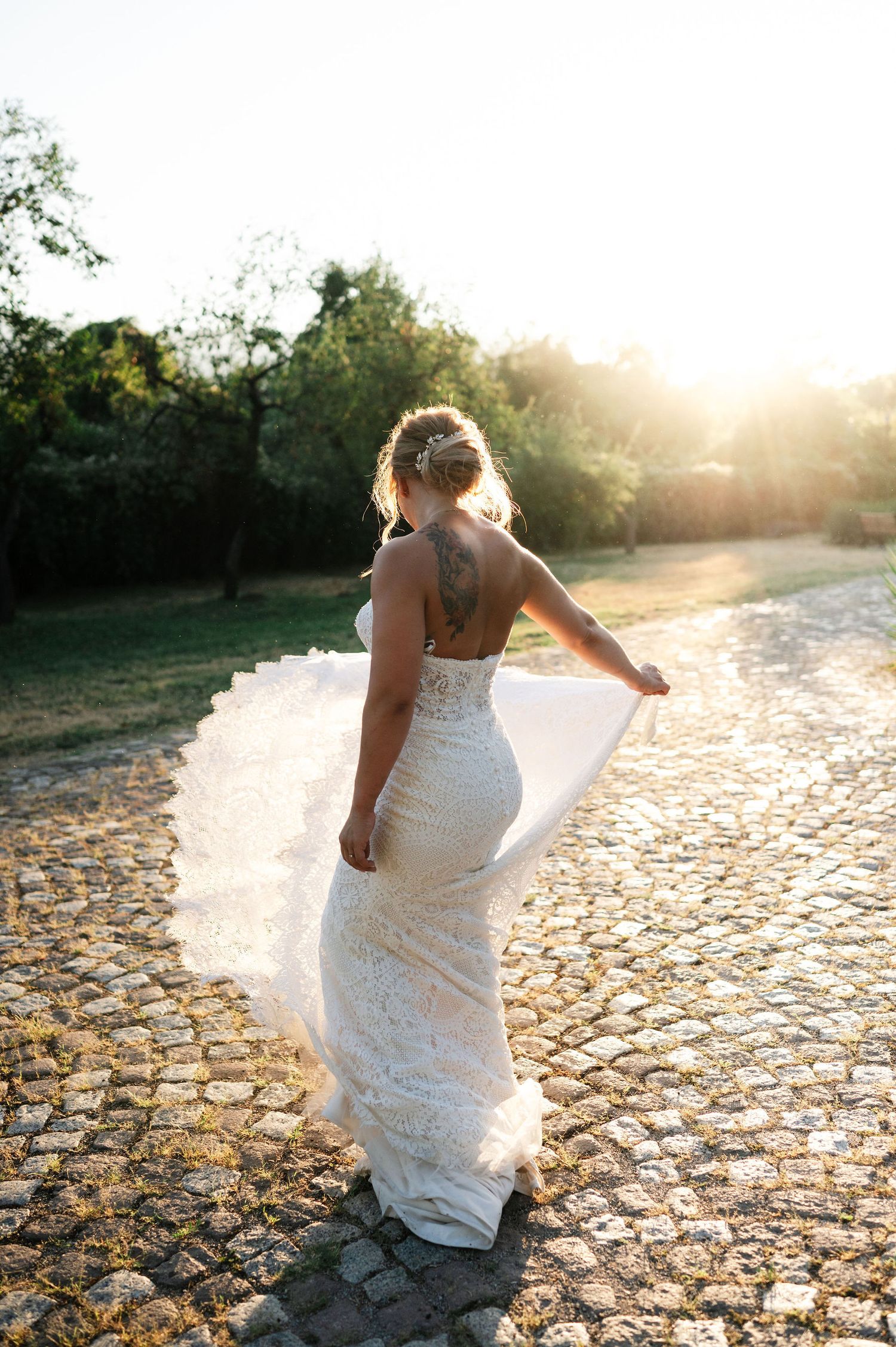 Wedding dress detail shot with intricate lace catching golden evening light on stone path.