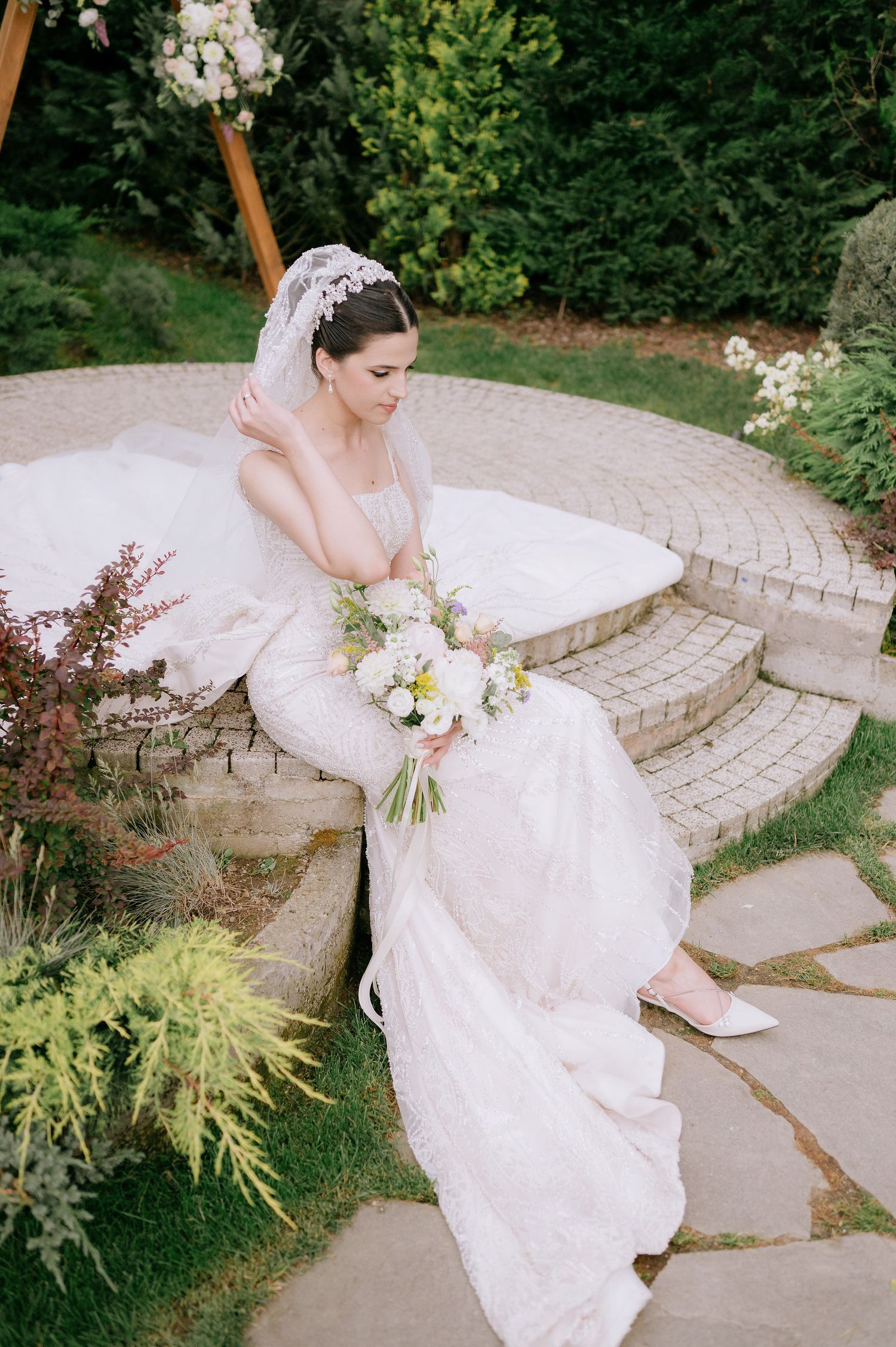 Elegant bridal portrait on curved stone steps with lush garden landscaping in background.