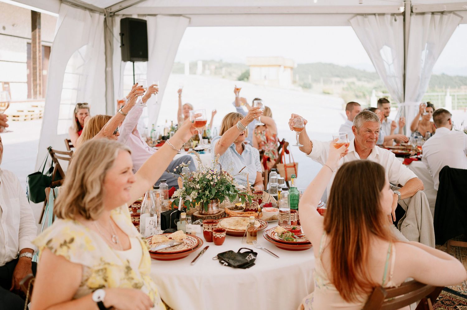 Wedding guests celebrate with raised arms at decorated tables under an outdoor tent.