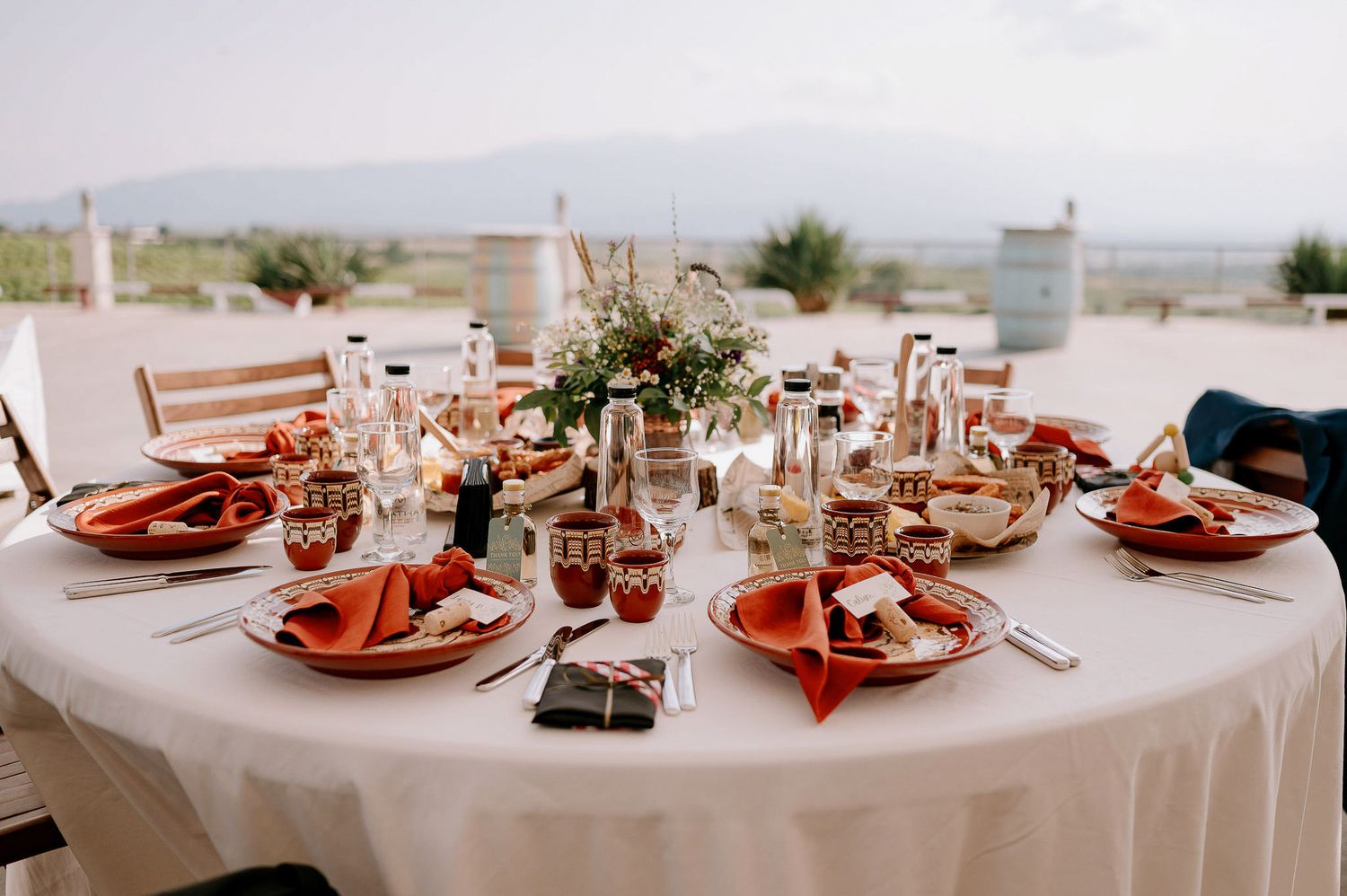 A formal table setting with coral napkins, glassware and floral centerpieces on a round white tablecloth.