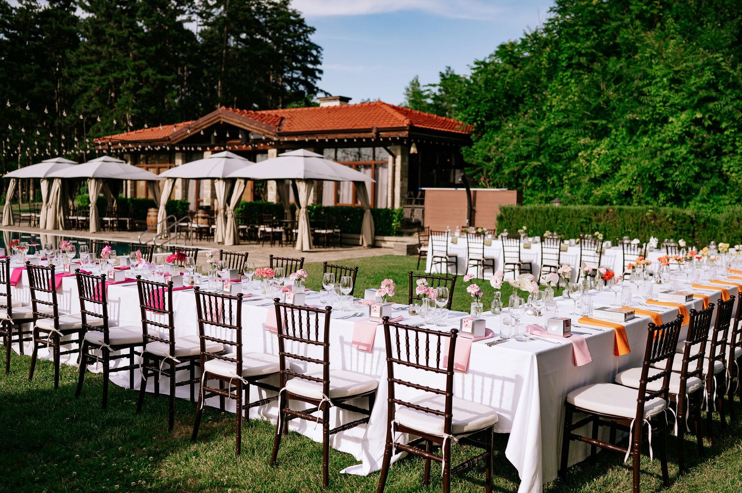 Elegant outdoor wedding reception setup with long tables, white linens and dark wooden chairs beside a covered pavilion.
