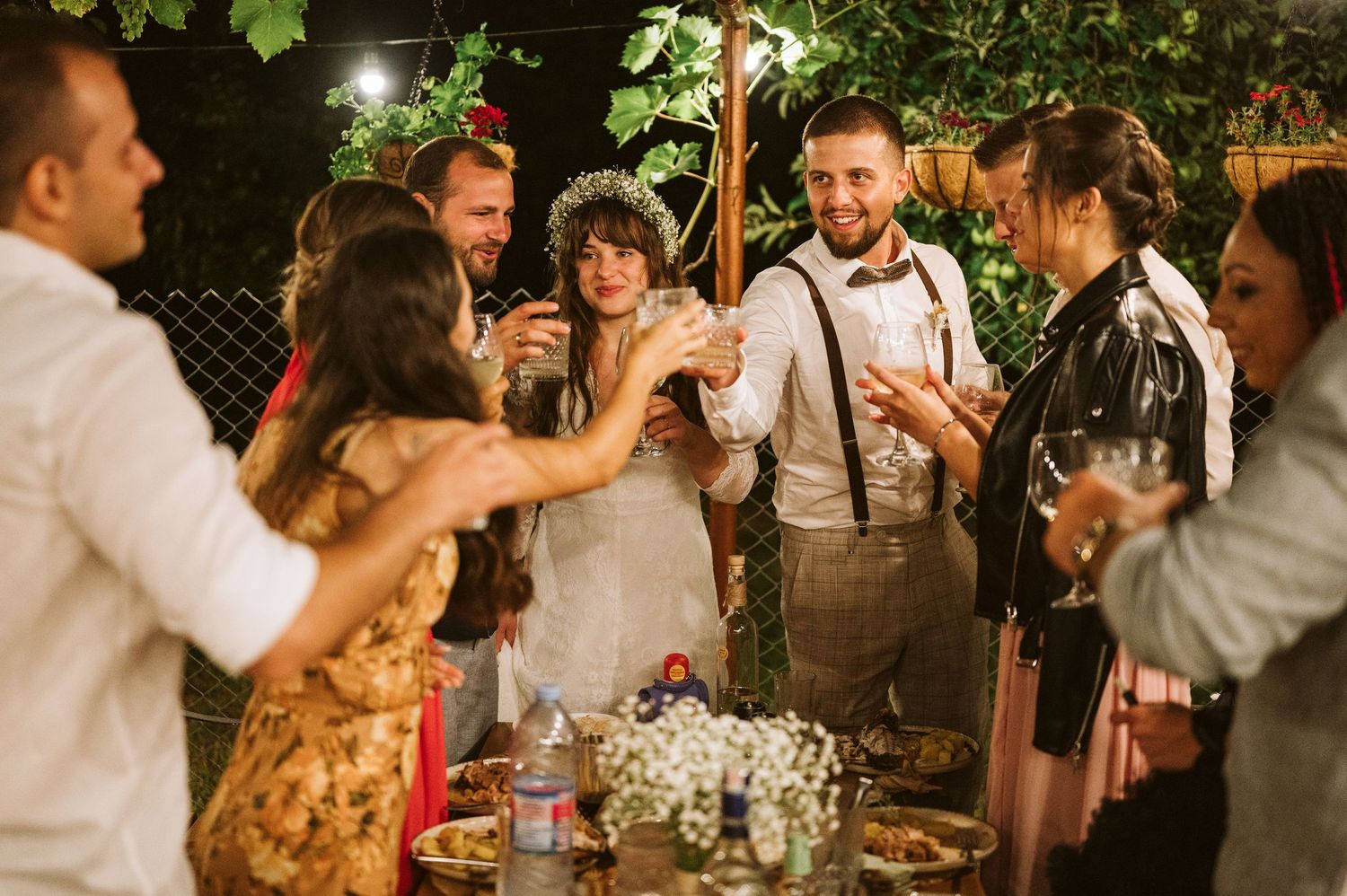 Wedding party enjoying a lively celebration under twinkling lights and garden foliage.