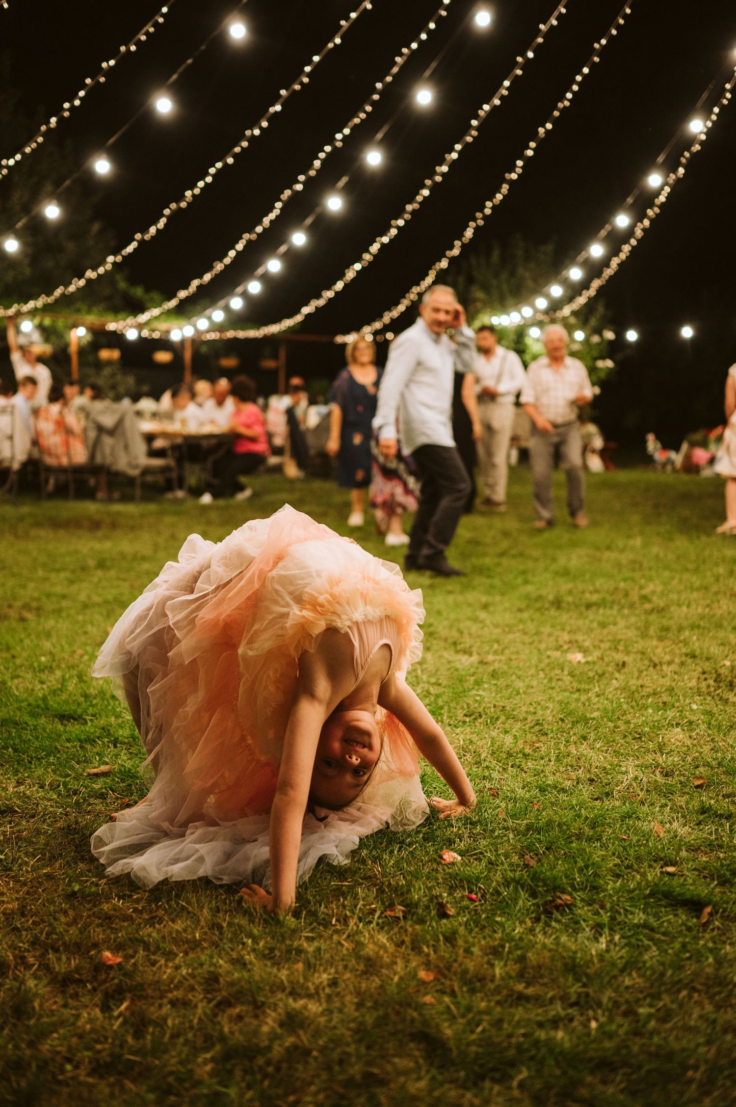 Child in pink tulle dress plays under magical string light canopy at outdoor reception.