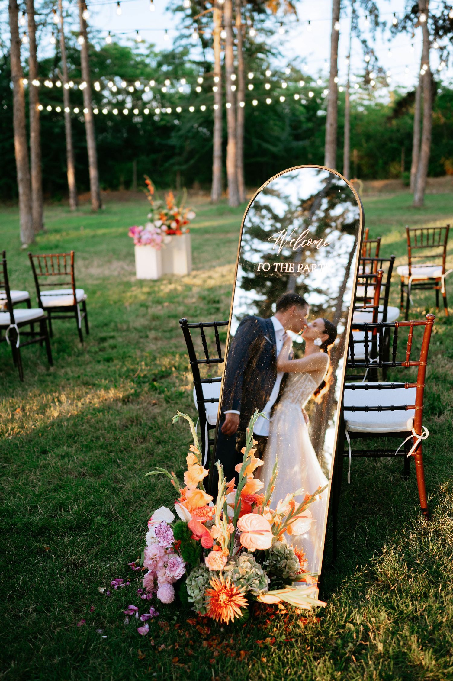 Outdoor wedding setup with string lights, wooden chairs, and floral arrangements in a wooded setting.
