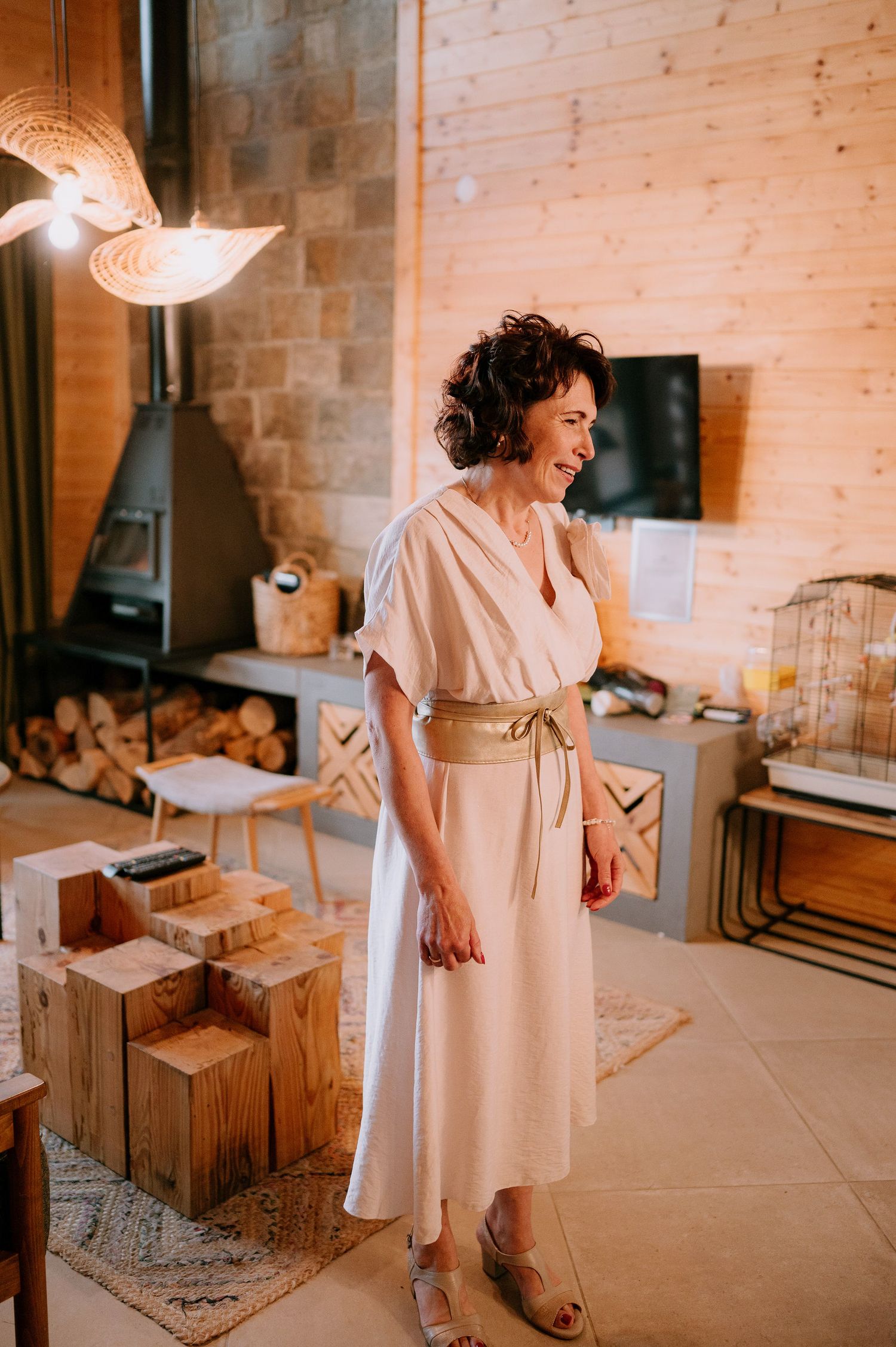 A person in a flowing beige dress stands in a rustic interior space with exposed brick walls.