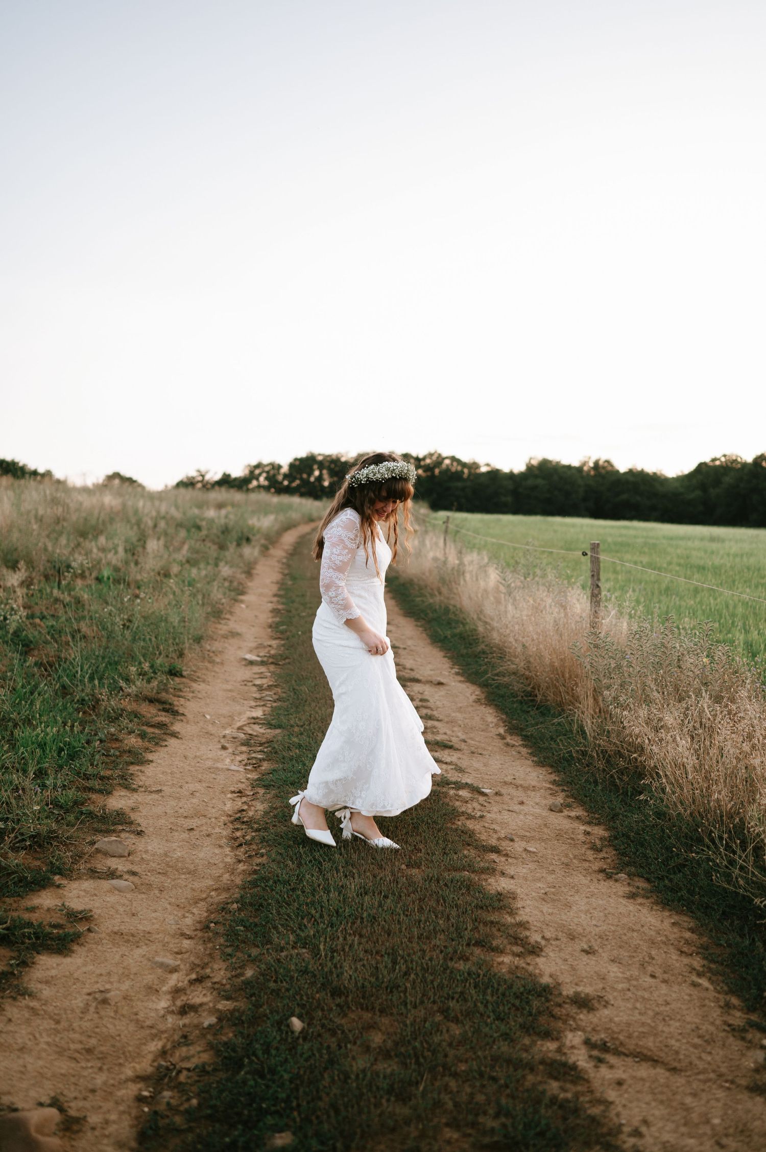 A bride in a lace dress walks along a dirt path through tall grass.