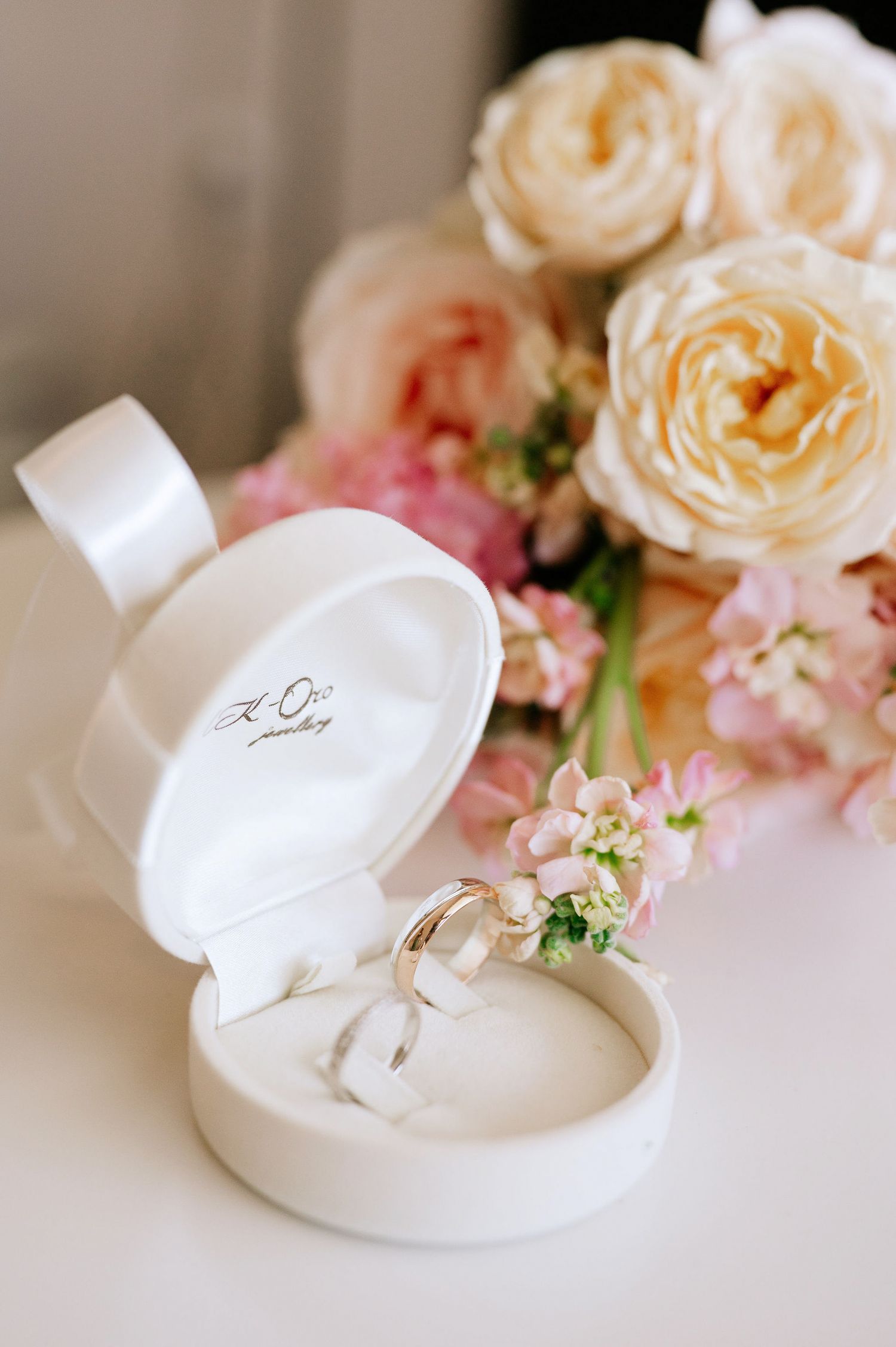 Wedding rings nestled in a white box surrounded by delicate peach roses and pink flowers.