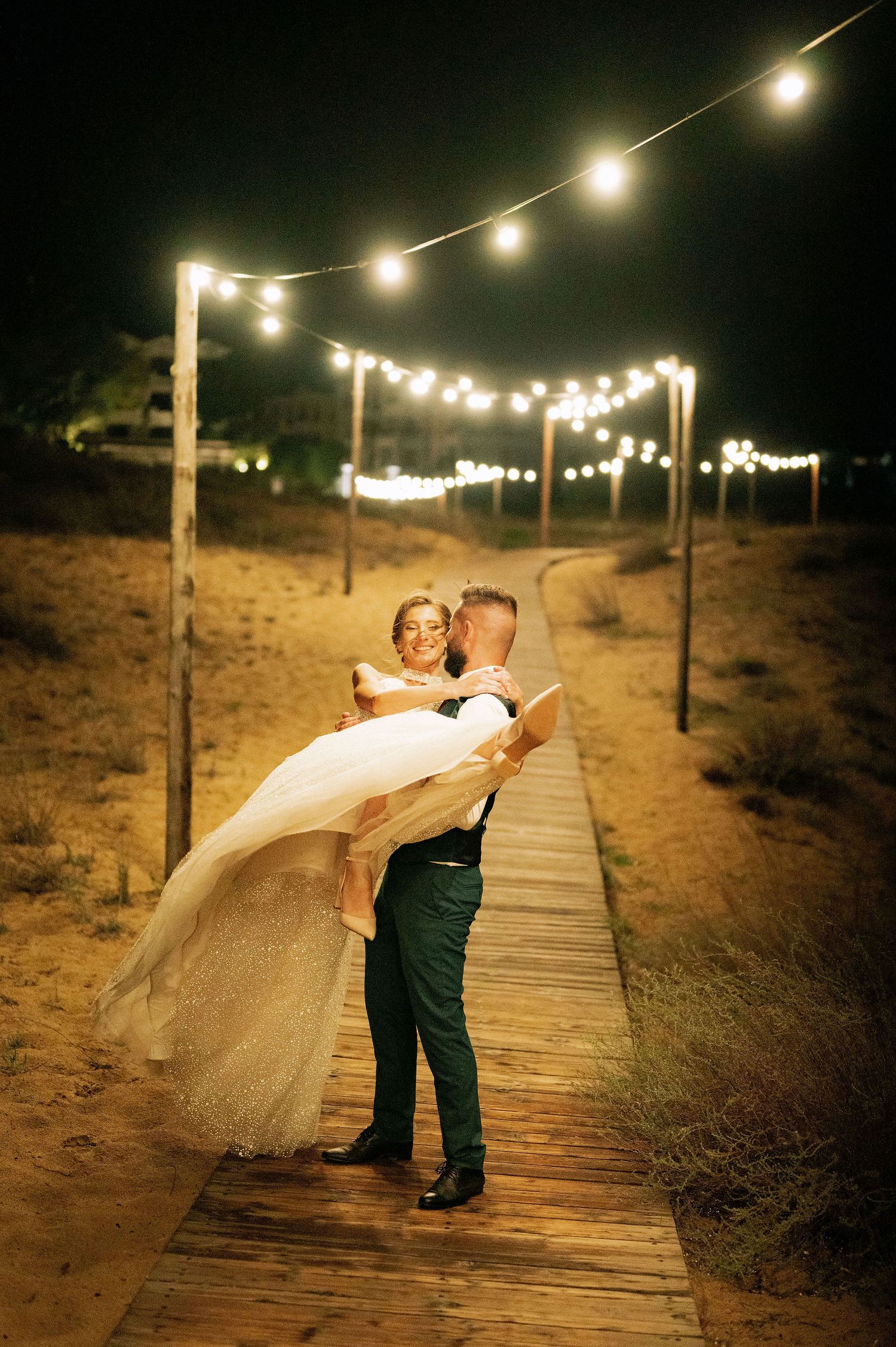 Couple sharing romantic dance moment on wooden boardwalk under string lights.