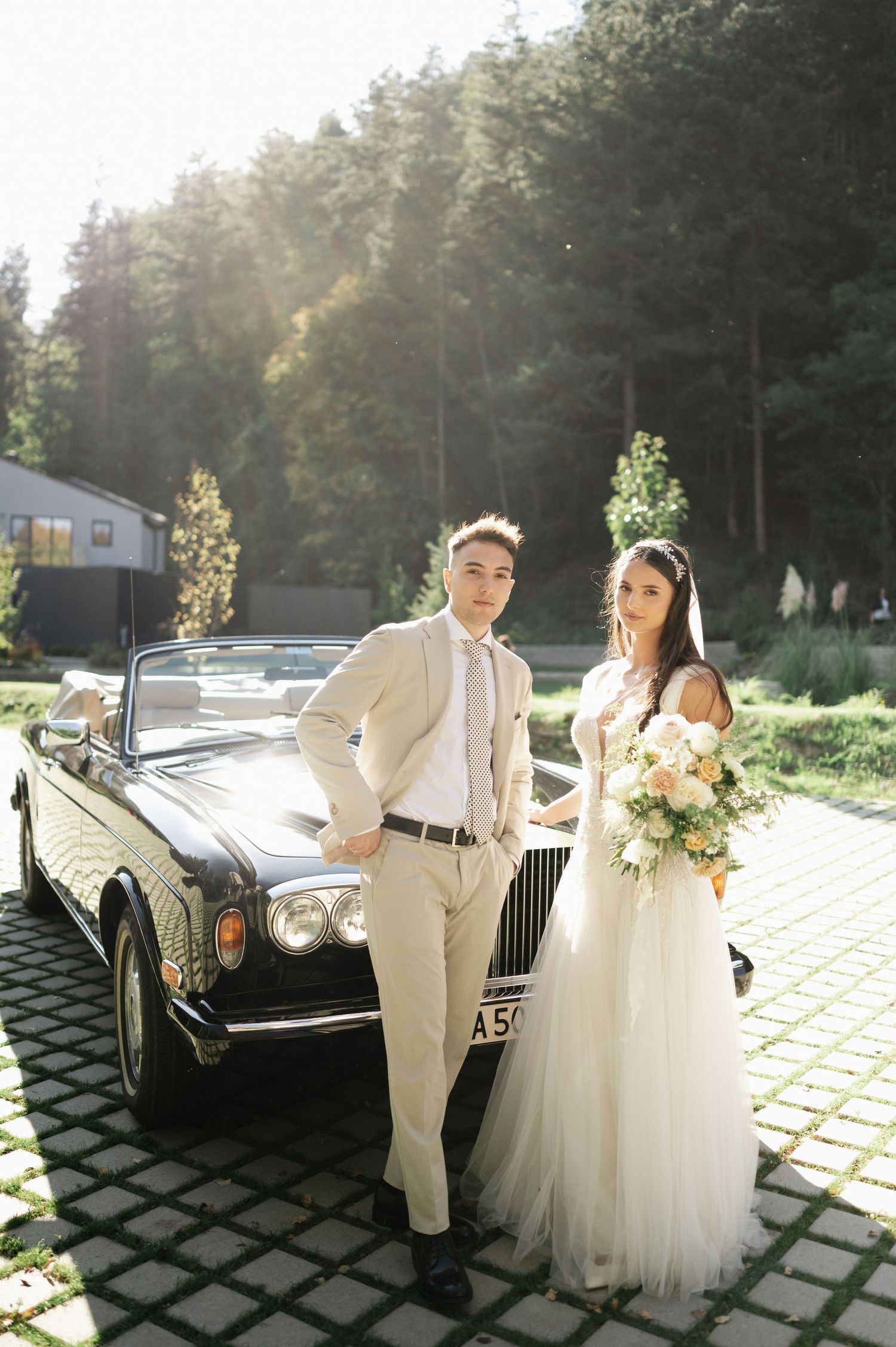 A wedding couple poses with a vintage black Rolls Royce convertible in a sunlit forest setting.