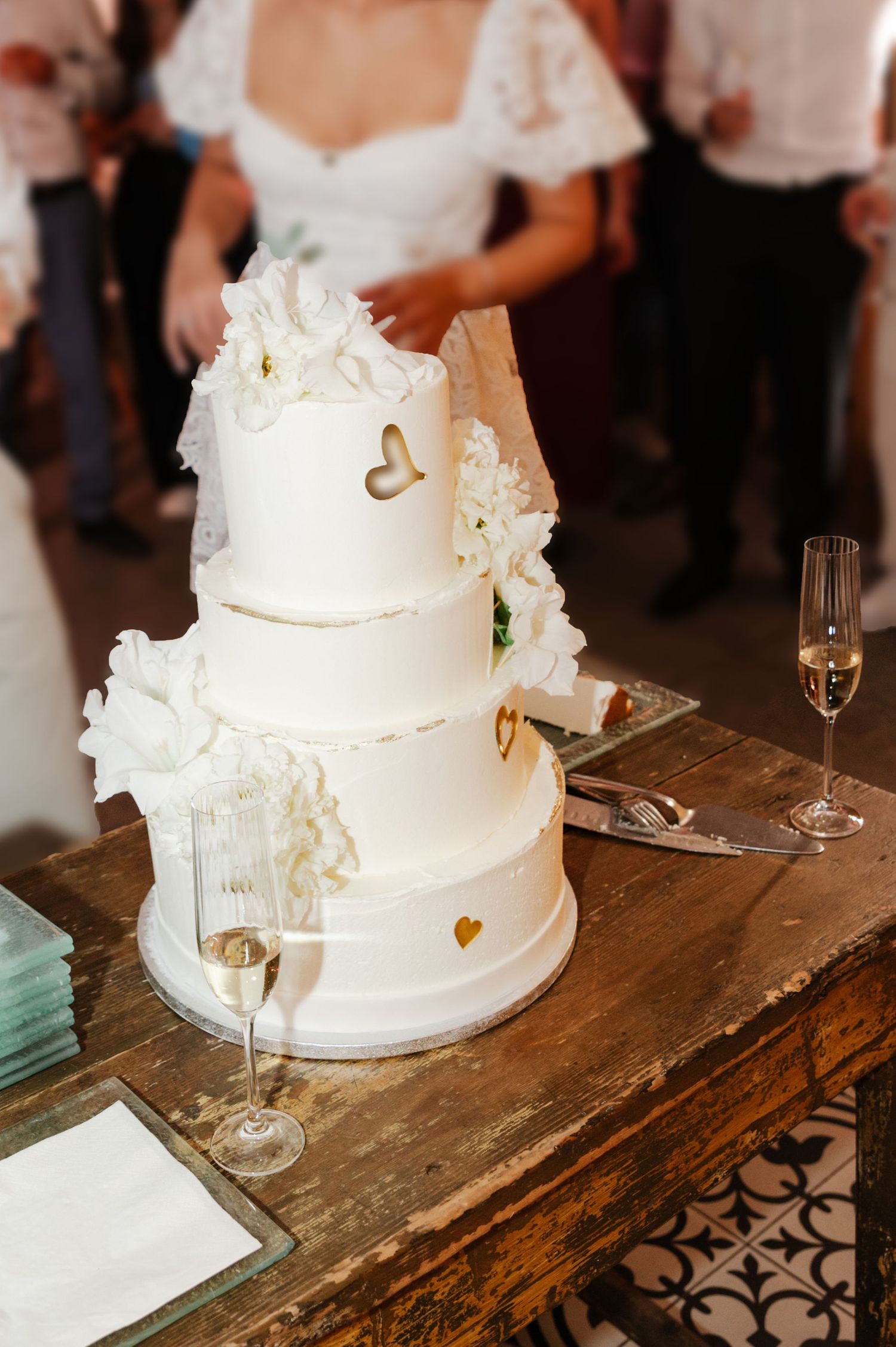 White tiered wedding cake with gold hearts displayed on rustic wooden table at reception.