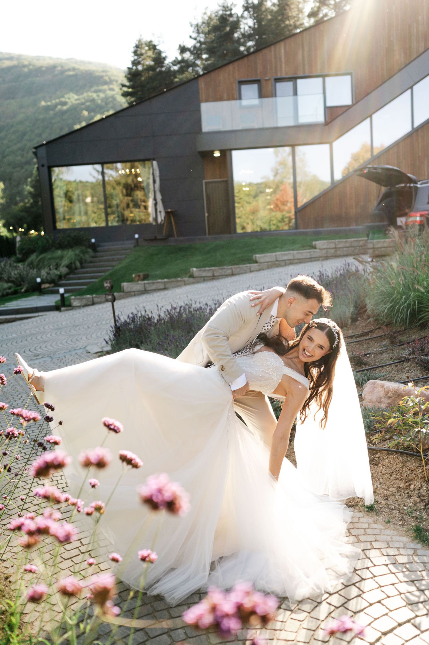 Wedding couple poses playfully on stone patio with pink flowers and modern building backdrop.