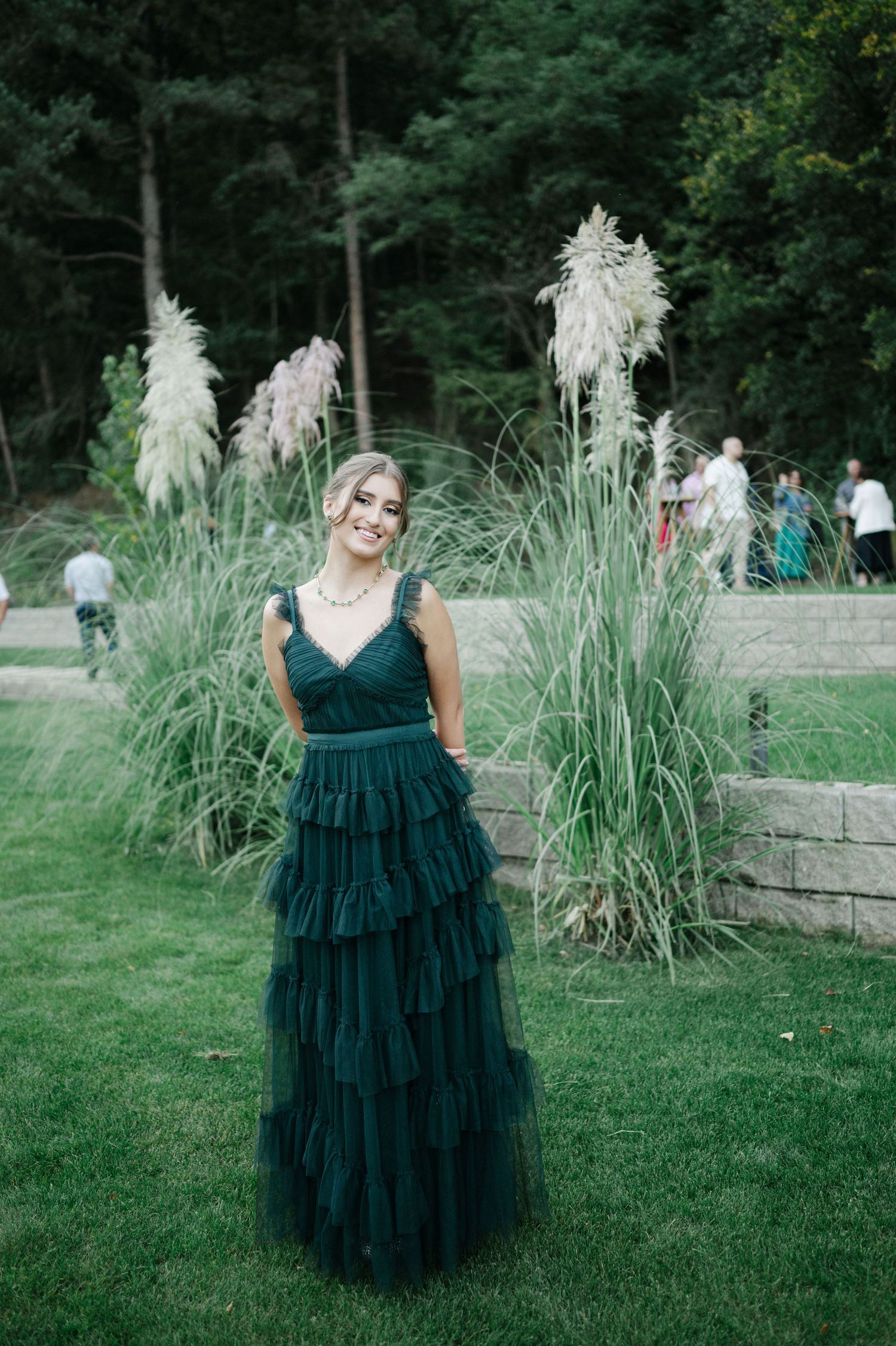 Person in an emerald green tiered dress poses on a grassy lawn with ornamental grasses.