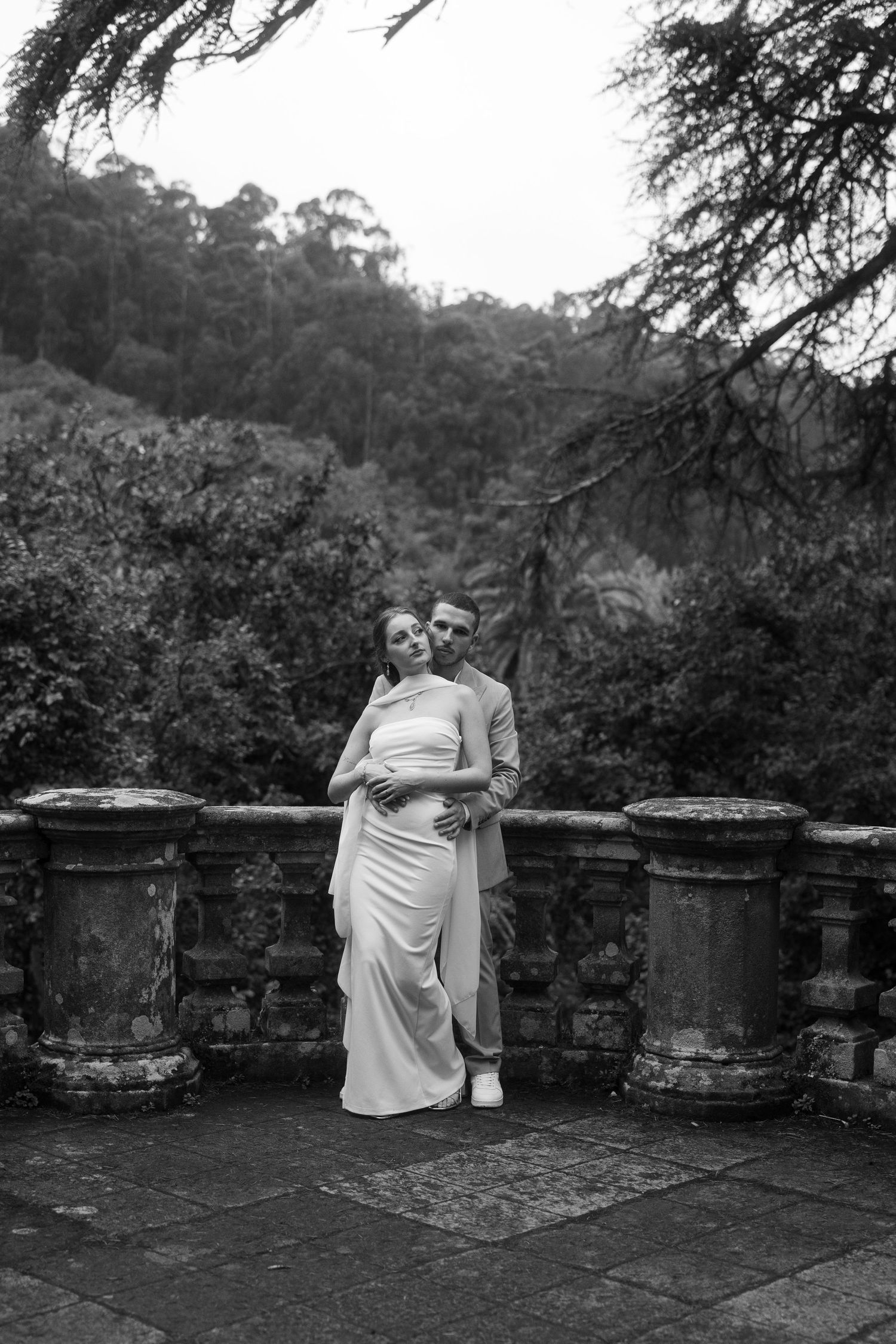 A couple in elegant attire sits together on a rustic stone terrace with trees in background.