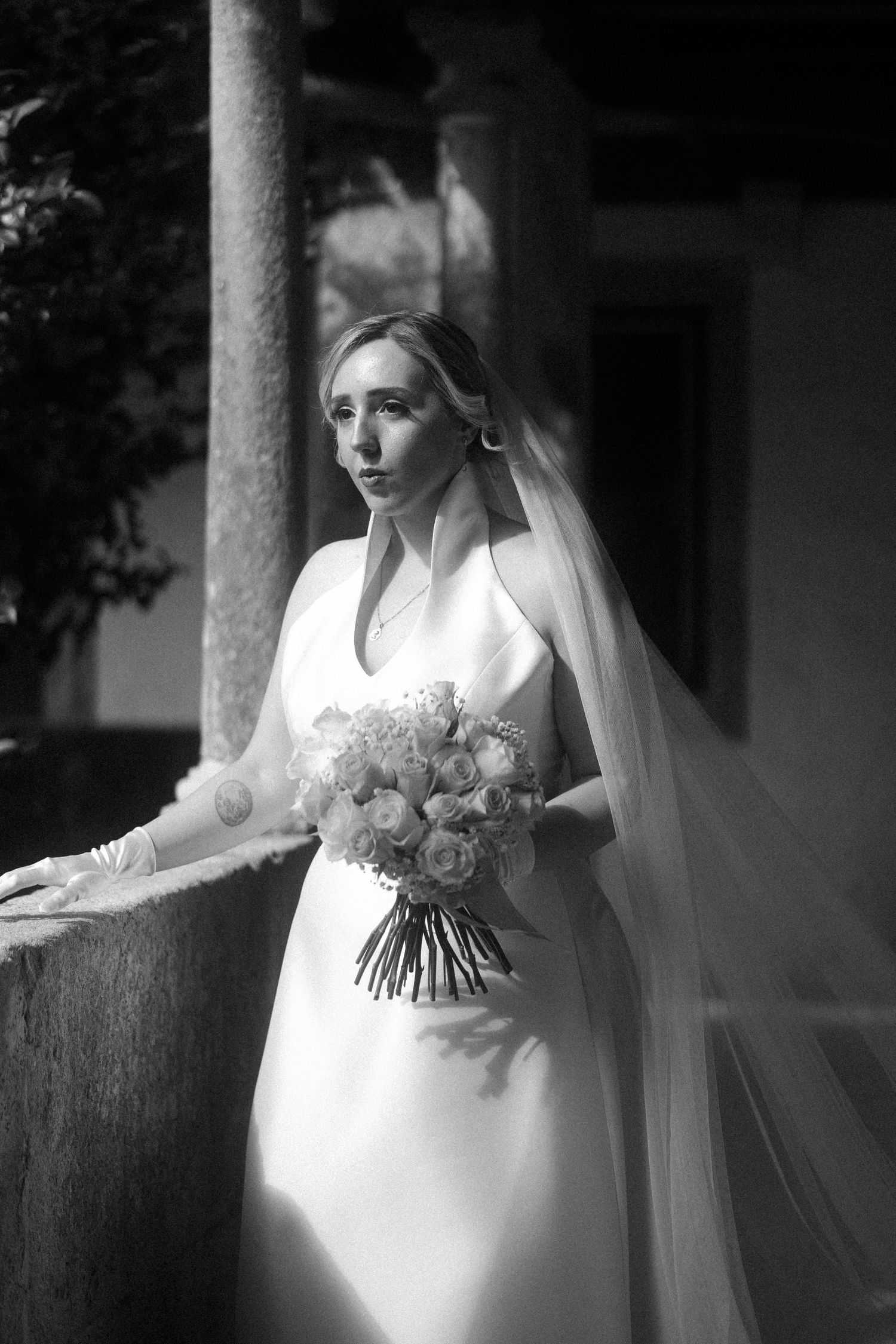 A black and white photo series of a bride in a flowing white gown holding a bouquet of flowers against a rustic backdrop.