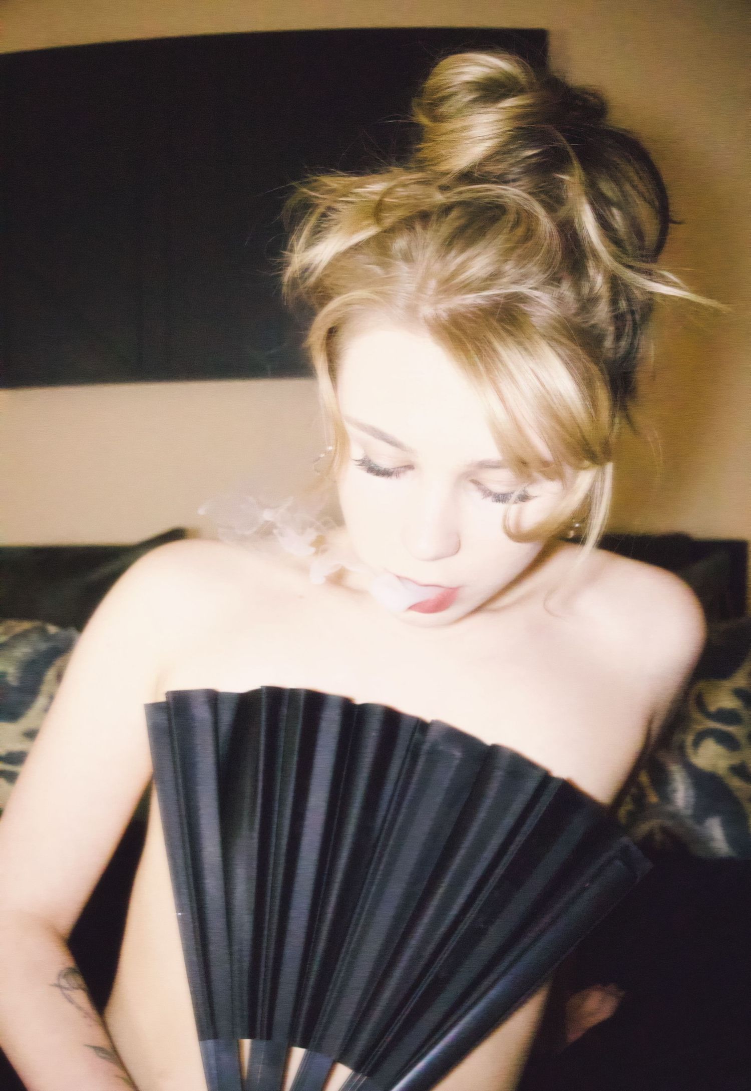 Blonde woman with messy bun hairstyle holding a black decorative fan, looking down gracefully.