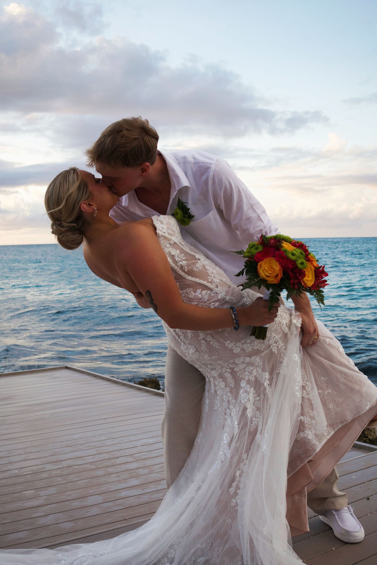 Bride and groom share a romantic dip kiss on a waterfront dock at sunset, bride holding colorful bouquet.