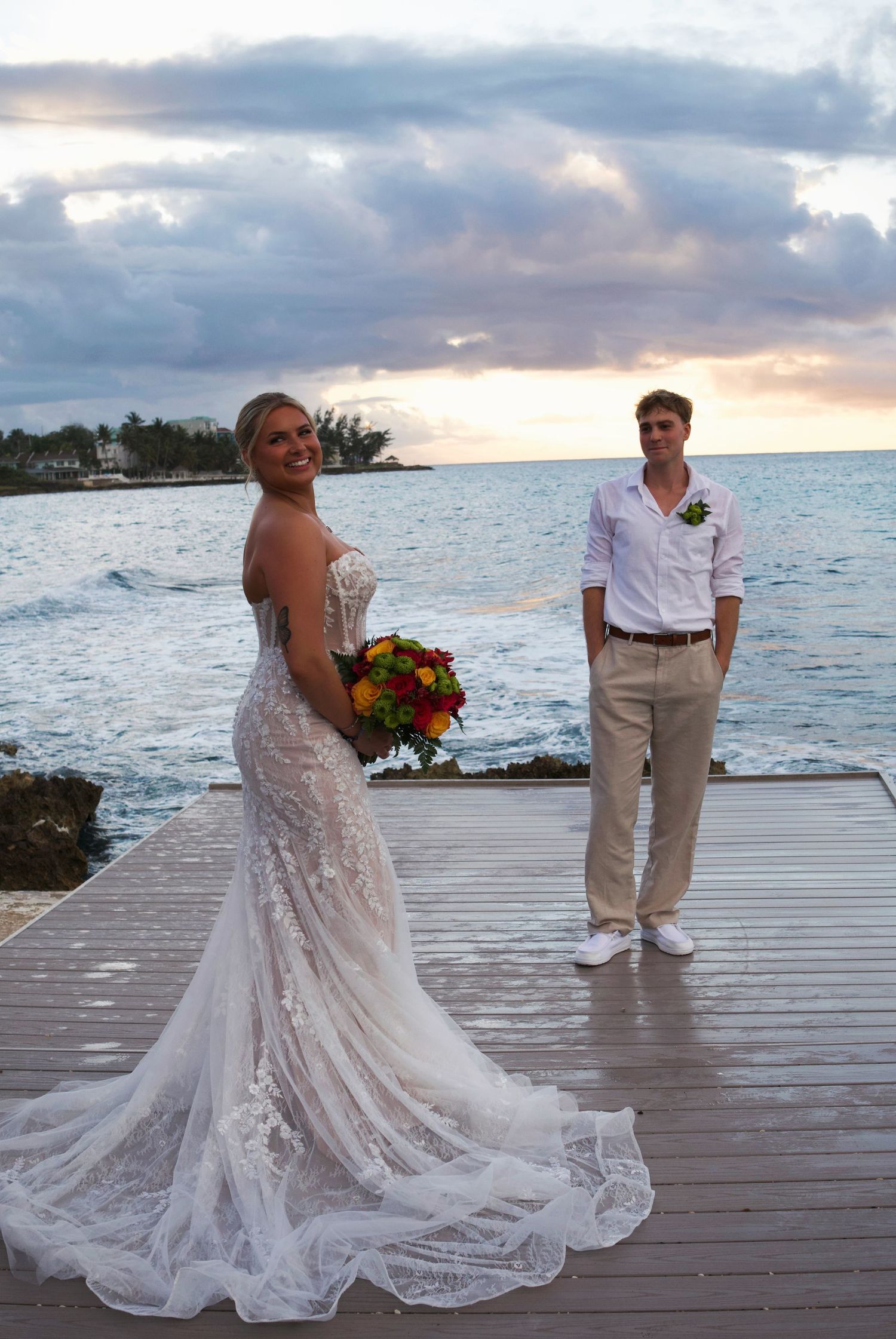 Bride in lace mermaid gown holds colorful bouquet on dock at sunset with groom in white shirt and khakis by ocean.