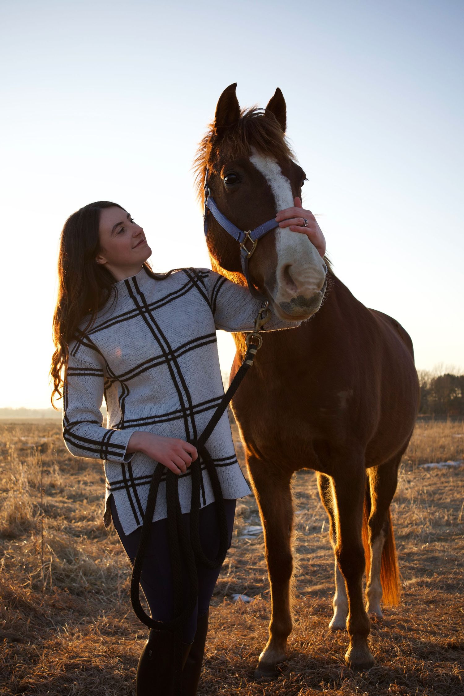Young woman in plaid sweater petting a brown horse with blaze marking in golden sunset light on open field.