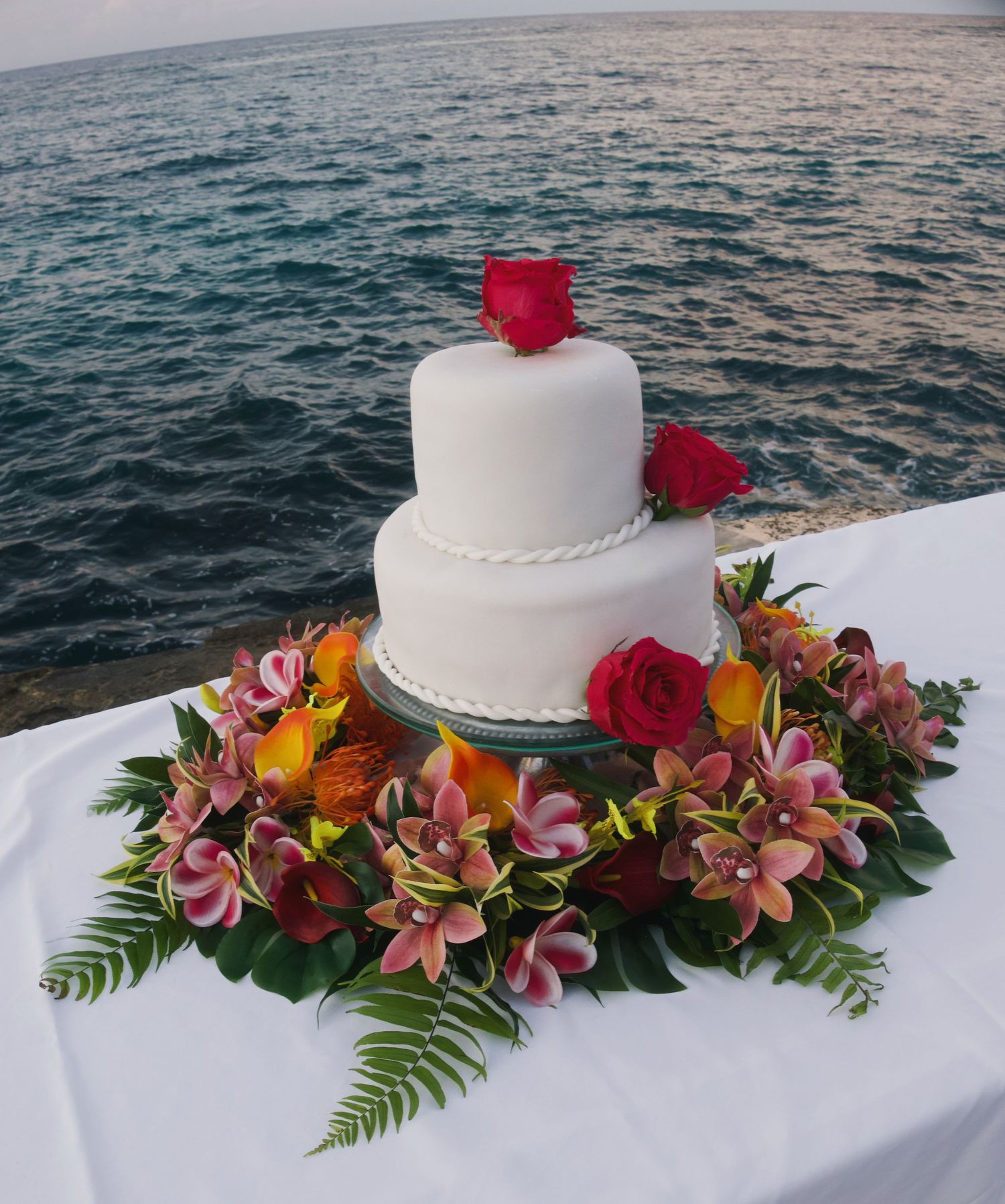 Two-tier white wedding cake adorned with red roses and tropical flowers on a table overlooking the ocean.