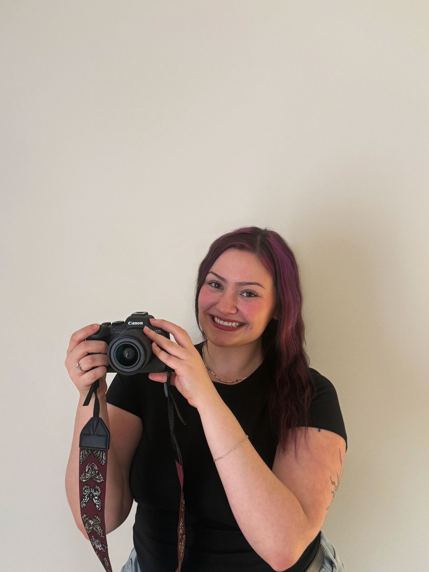 Woman with purple hair smiling and holding a Canon camera with a decorative strap against a neutral wall.