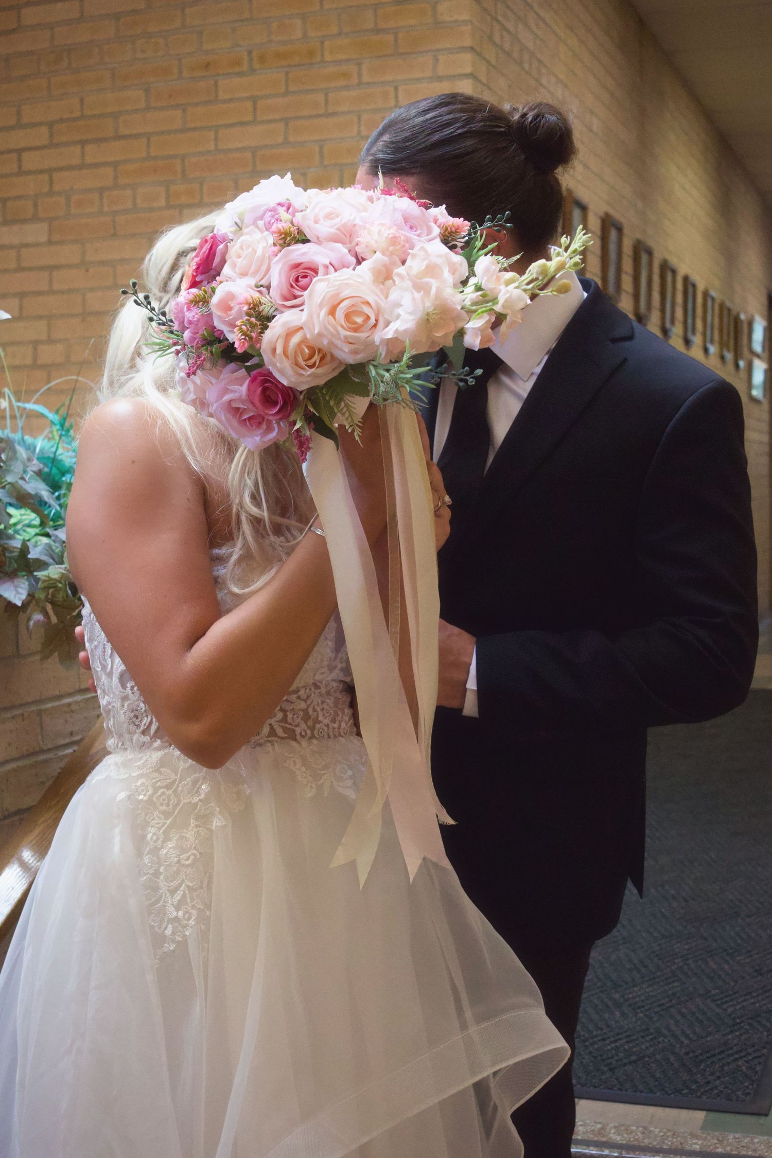 Bride and groom kissing behind a pink rose bouquet in wedding attire near a brick wall.