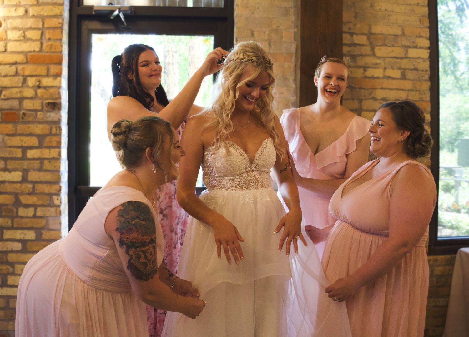 Bride in white gown surrounded by smiling bridesmaids in pink dresses getting ready in a rustic venue.