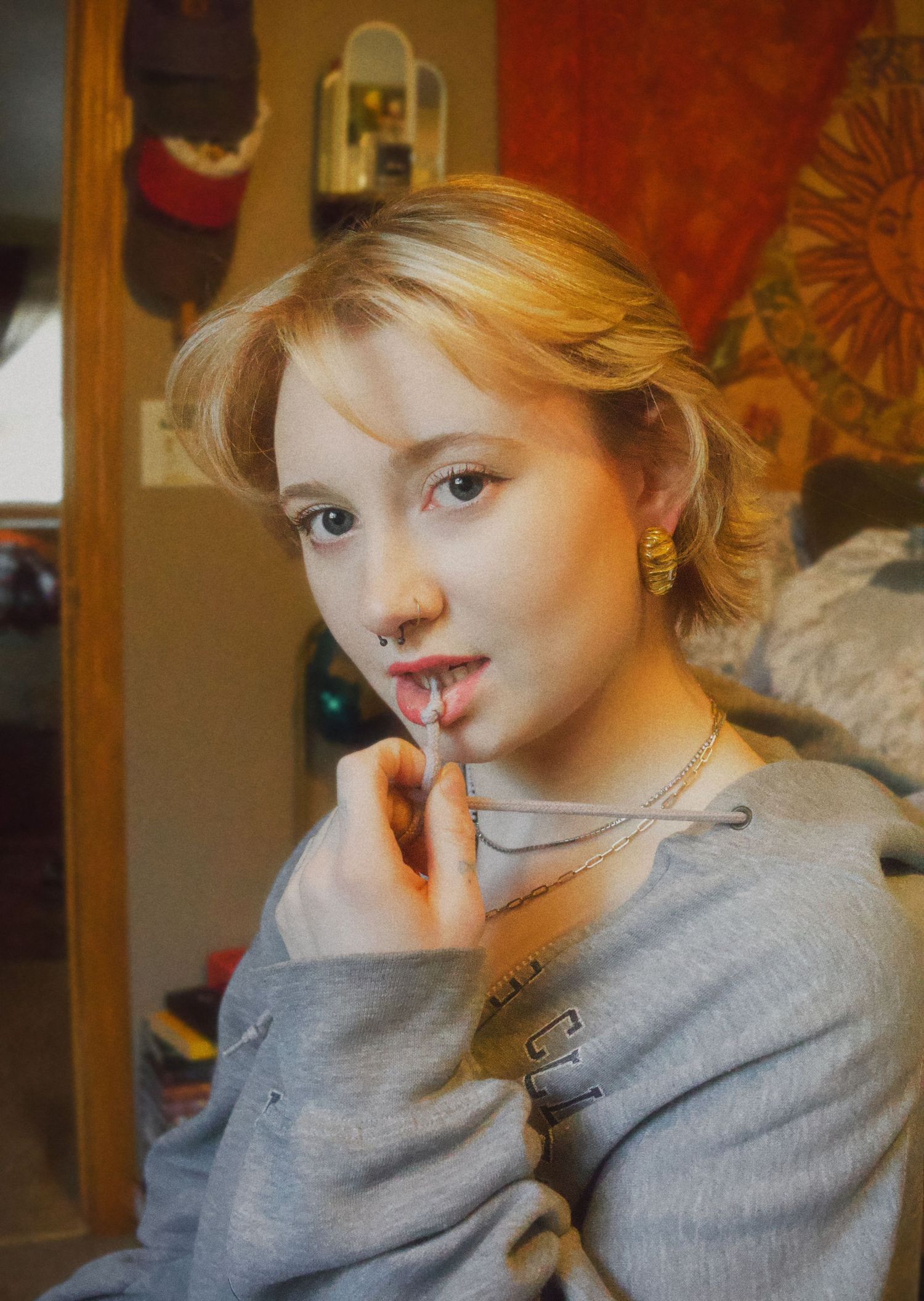 Young woman with short blonde hair wearing a gray sweater and gold earrings, posing indoors with hand near her lips.