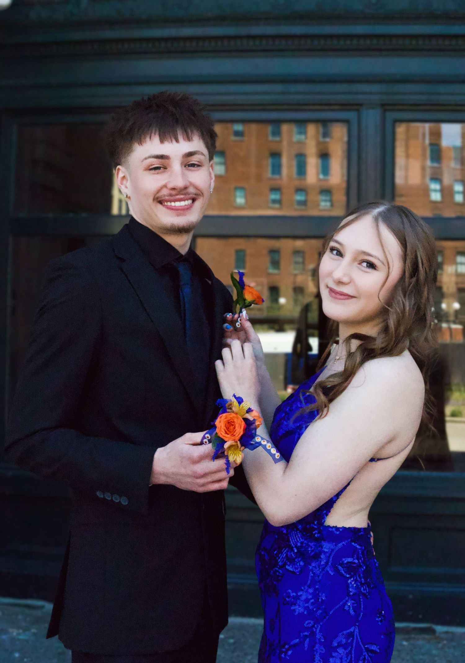 A smiling couple in prom attire, man in black suit and woman in royal blue gown, posing with orange corsages outdoors.