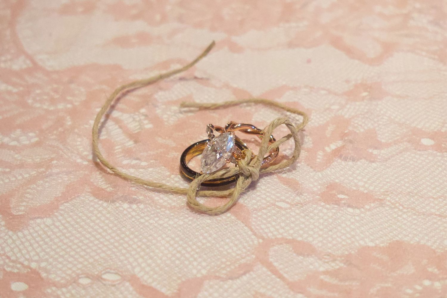 Gold and diamond engagement ring with wedding bands tied together with twine on pink lace fabric.