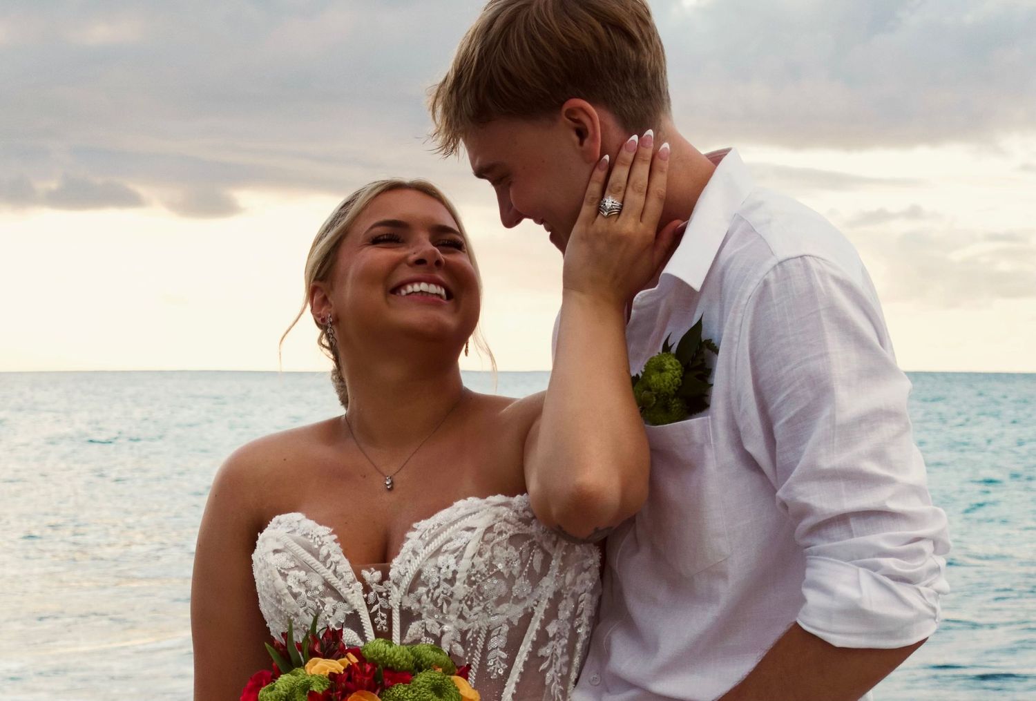 Bride and groom share a romantic beach moment at sunset, with colorful bouquet and ocean backdrop.