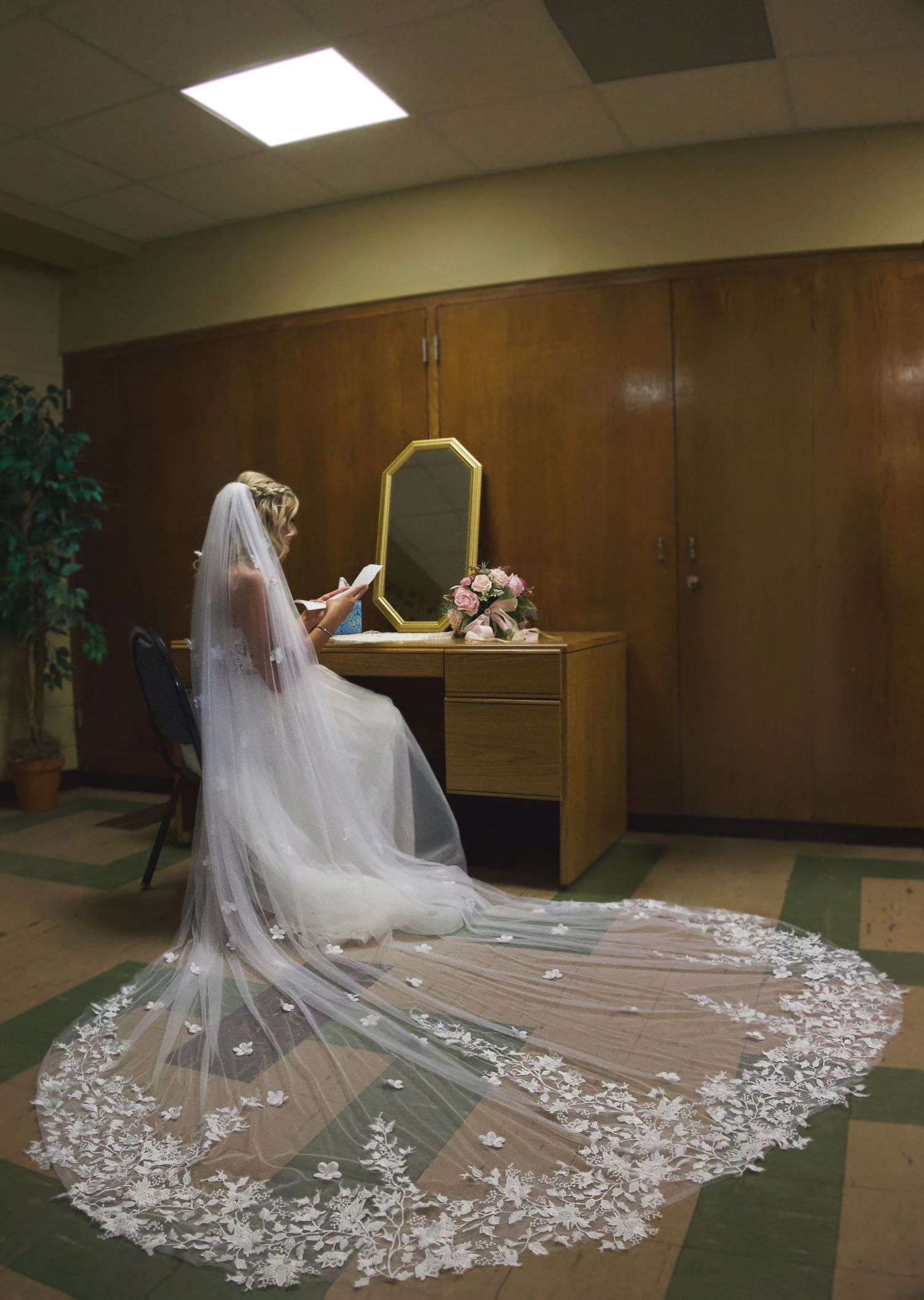 Bride in white gown with long lace cathedral veil seated at vanity mirror, holding bouquet of pink flowers.