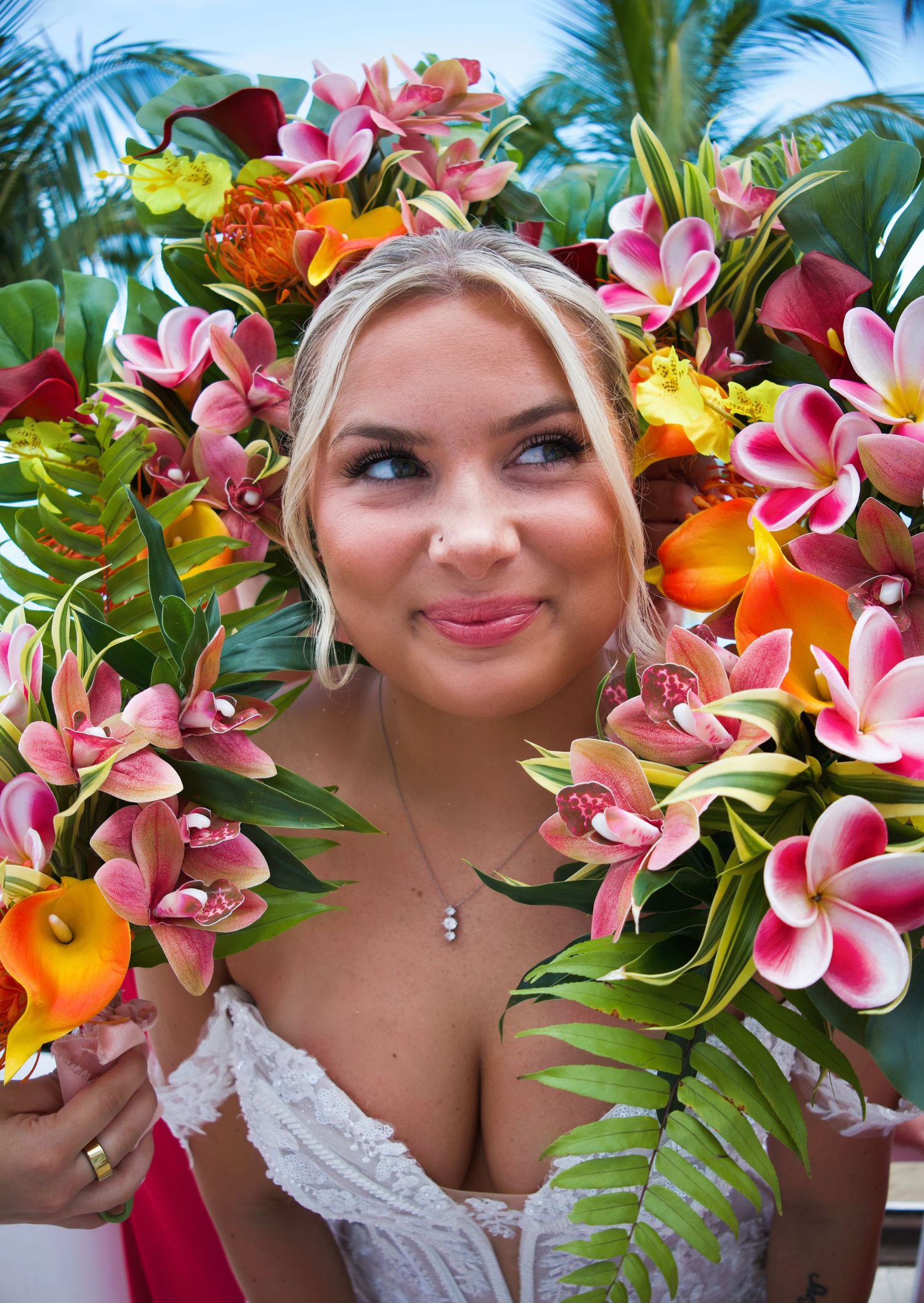 Smiling blonde bride holding a vibrant tropical bouquet of pink plumeria, orchids, and calla lilies outdoors.