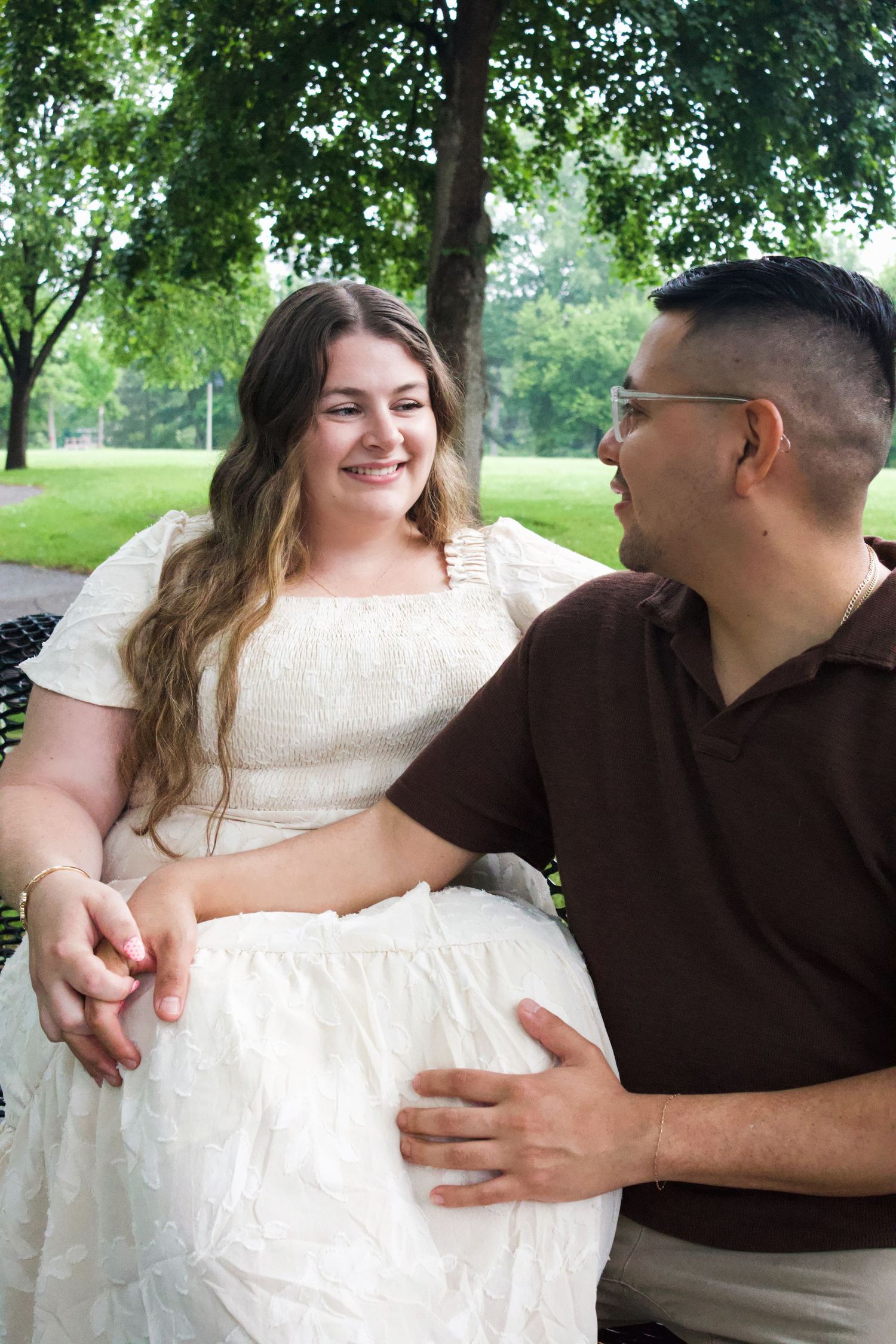 A smiling couple sits on a park bench, woman in white dress holding hands with man in brown shirt.