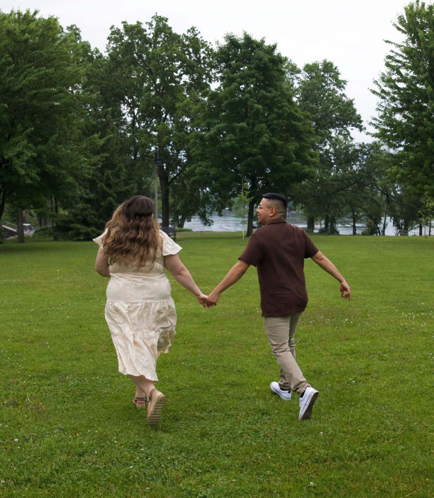 Couple holding hands running through a lush green park with tall trees and a lake in the background.