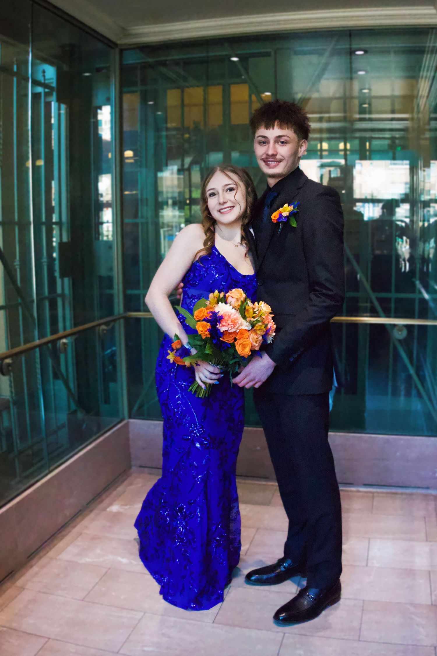 A young couple dressed for prom, she in a blue sequin gown holding orange flowers, he in a black suit with boutonniere.
