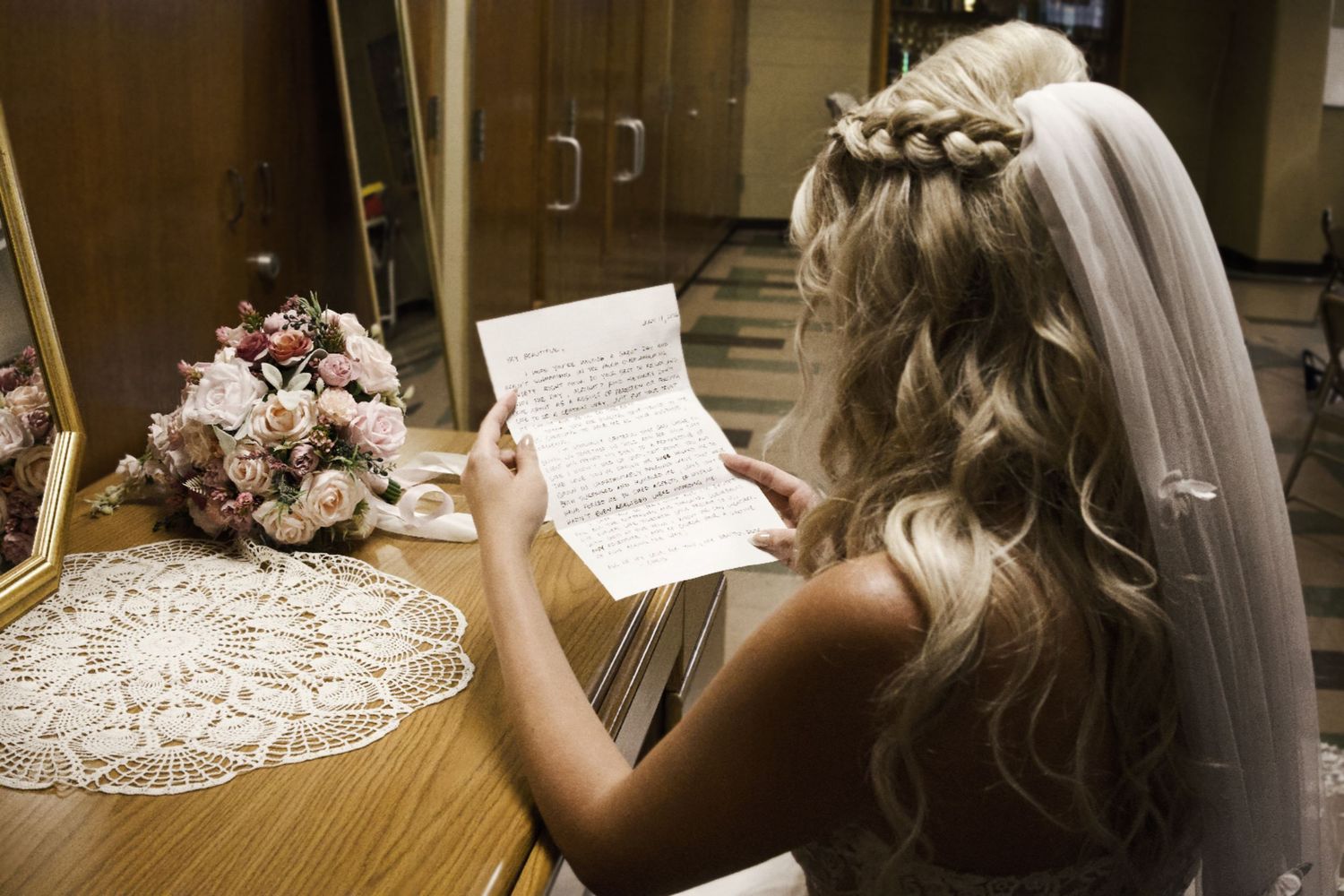 Bride in wedding veil reads a heartfelt letter beside a floral bouquet, reflected in a mirror.