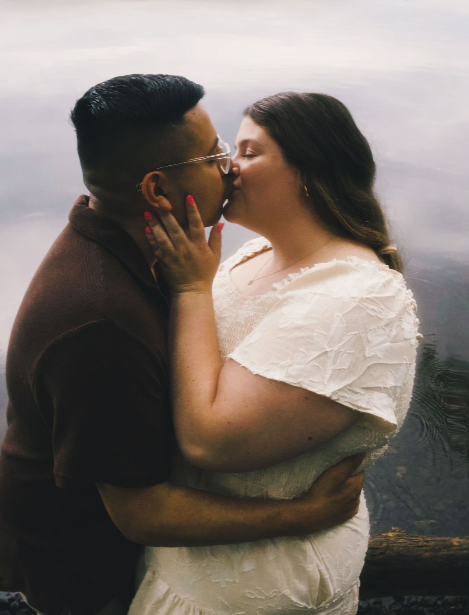 Couple sharing a romantic kiss outdoors near water, woman in white dress and man in brown shirt.